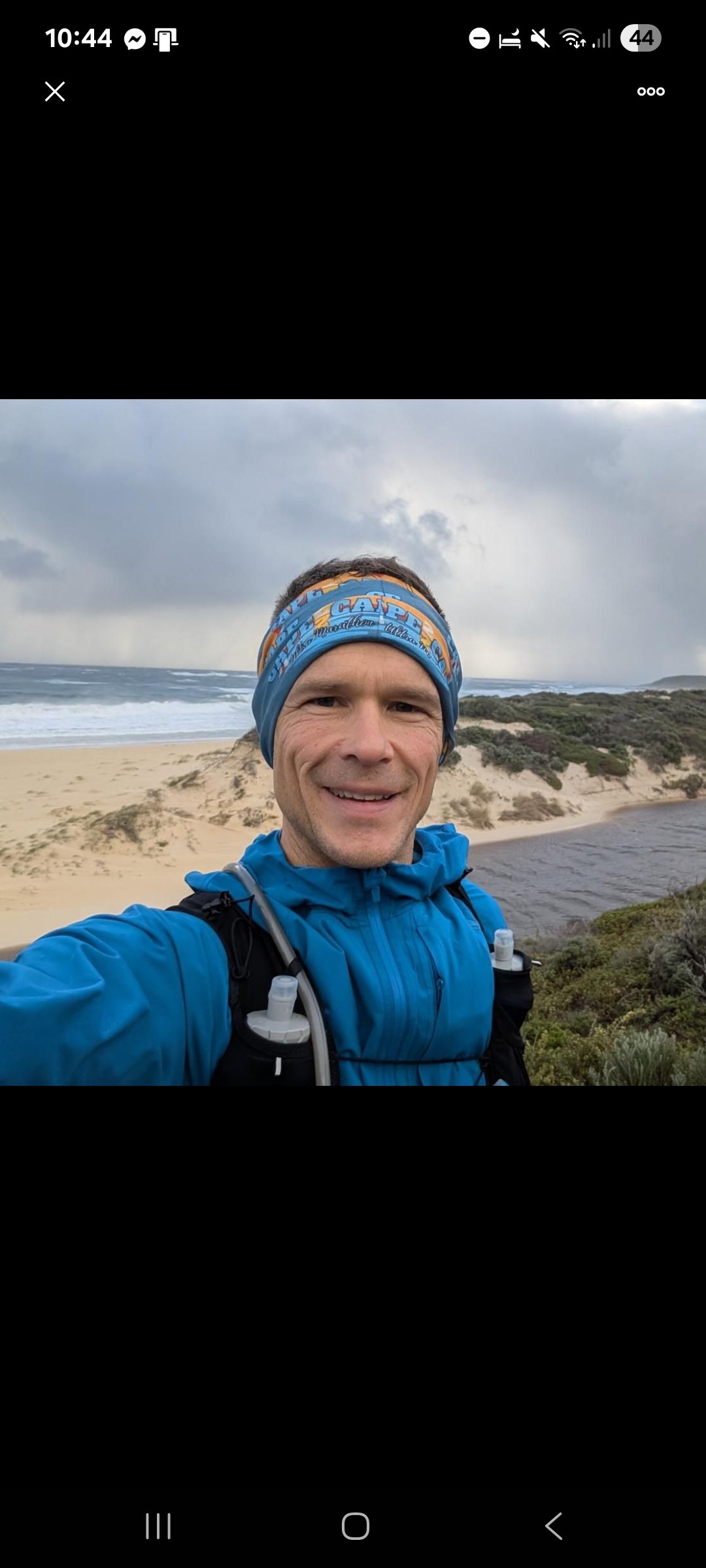 A man taking a selfie on a beach with sand dunes and ocean waves in the background, wearing a blue jacket and headband, carrying a backpack.