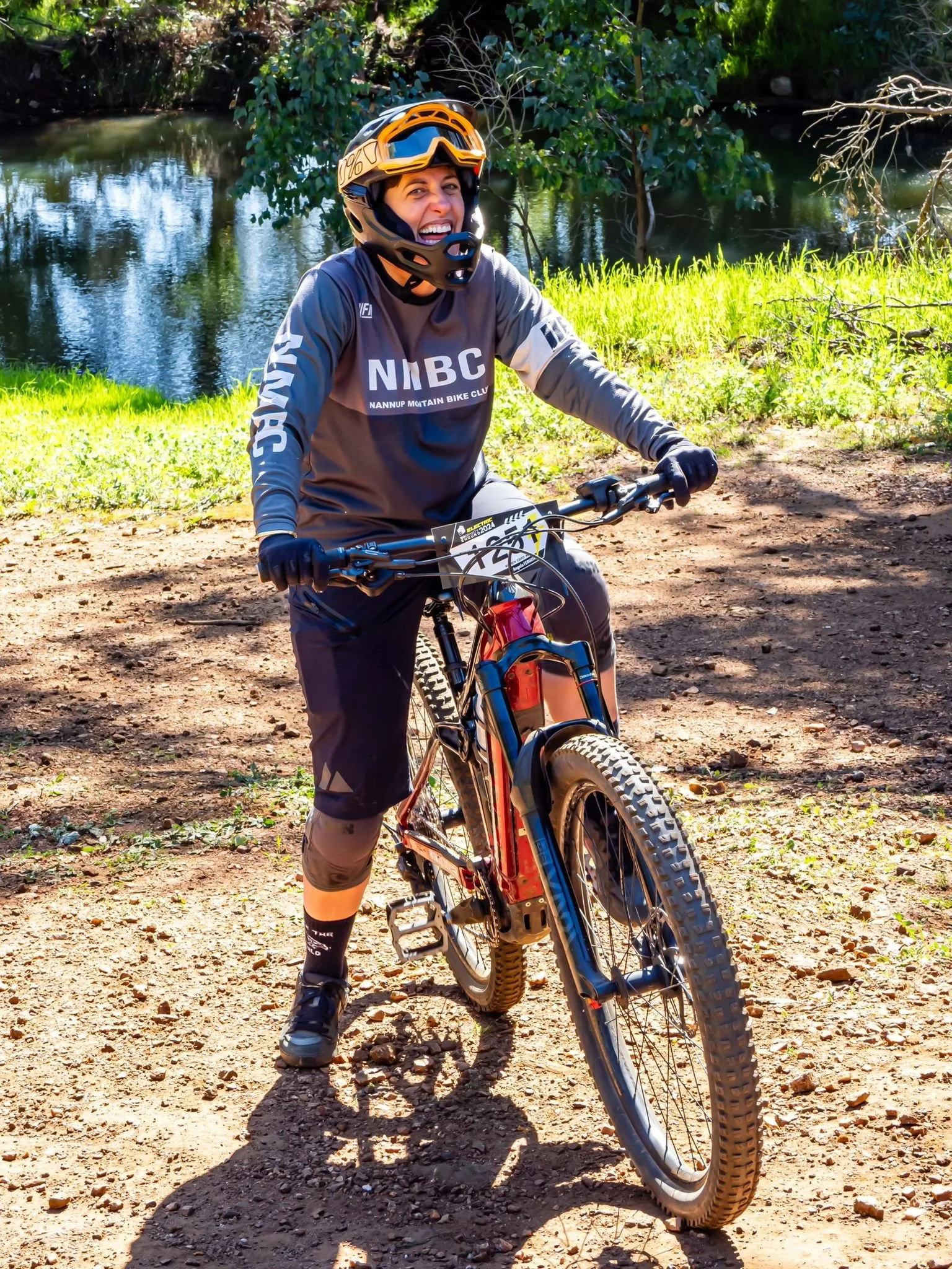 Woman in biking gear and helmet smiling while riding a mountain bike on a dirt trail near a body of water with trees and greenery in the background.