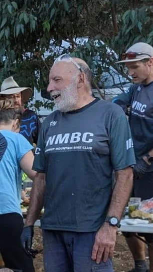 Older man with a white beard smiling, wearing a navy NMBC T-shirt, surrounded by people outdoors.