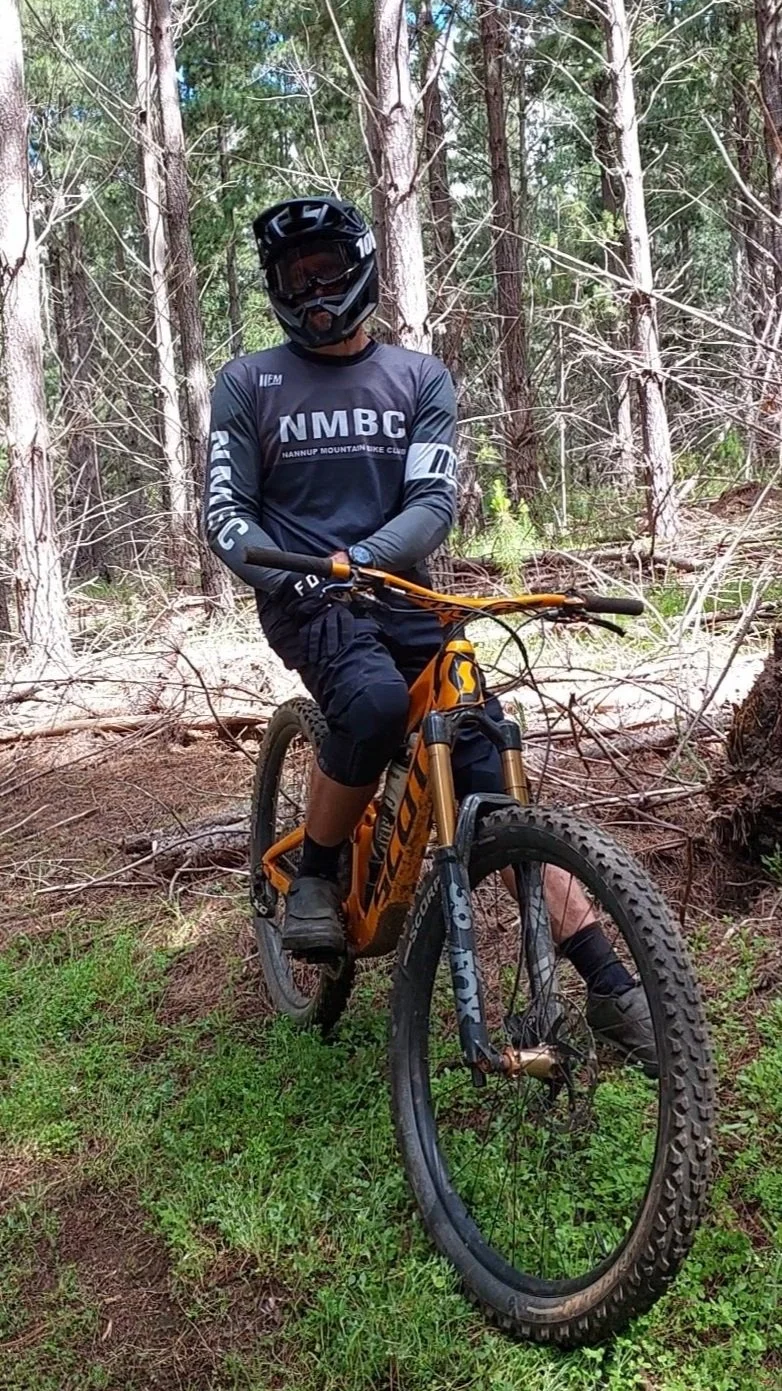 A mountain biker wearing a black full-face helmet, a NMBC long sleeve jersey, black shorts, and riding an orange mountain bike on a dirt trail through a forest with trees and grass.
