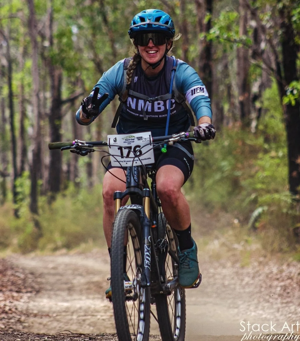 A woman riding a mountain bike on a trail in a forest, wearing a helmet, sunglasses, and a race bib, smiling and making a peace sign.