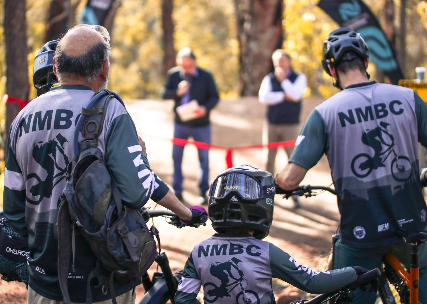 A group of people wearing biking gear with NMBC logos, preparing for or participating in a biking event in a wooded area, with some individuals standing and one person sitting on a mountain bike.