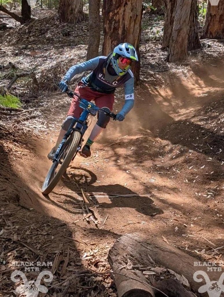 Mountain biker wearing a blue helmet, goggles, and protective gear riding on a dirt trail through a forest with trees and fallen logs.