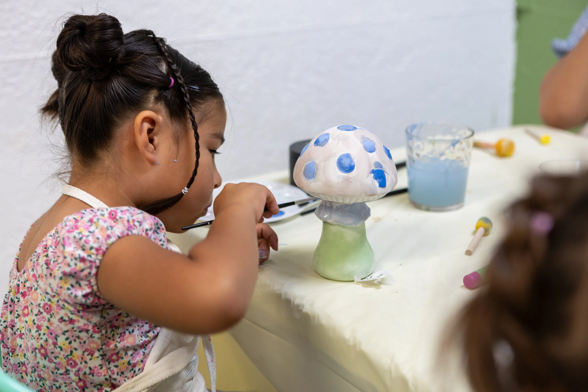 Close-up of a woman painting a butterfly on a white ceramic plate with a small paintbrush, sitting at a wooden table.