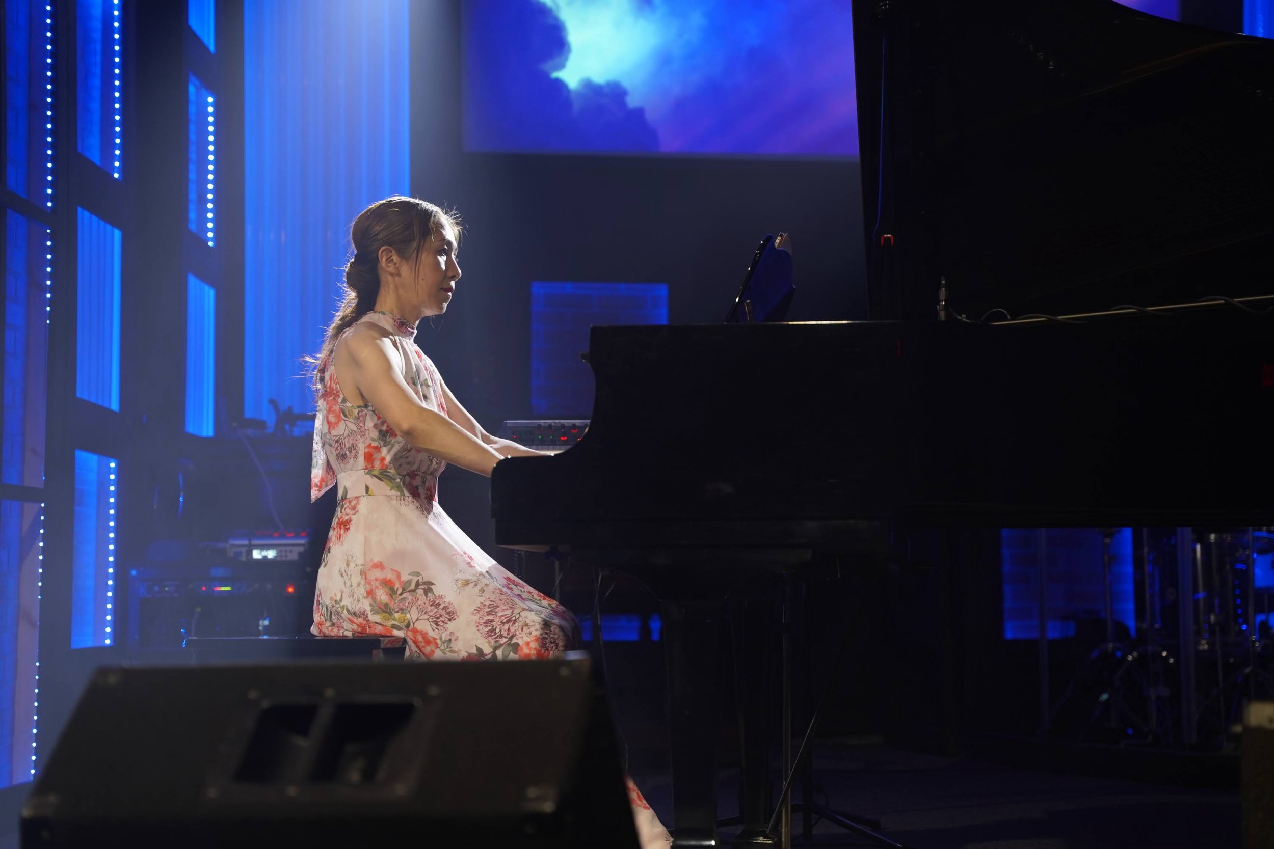 Nana Morimoto performing a classical piano solo on stage in a floral dress, playing a grand piano under blue lighting with a large screen in the background.