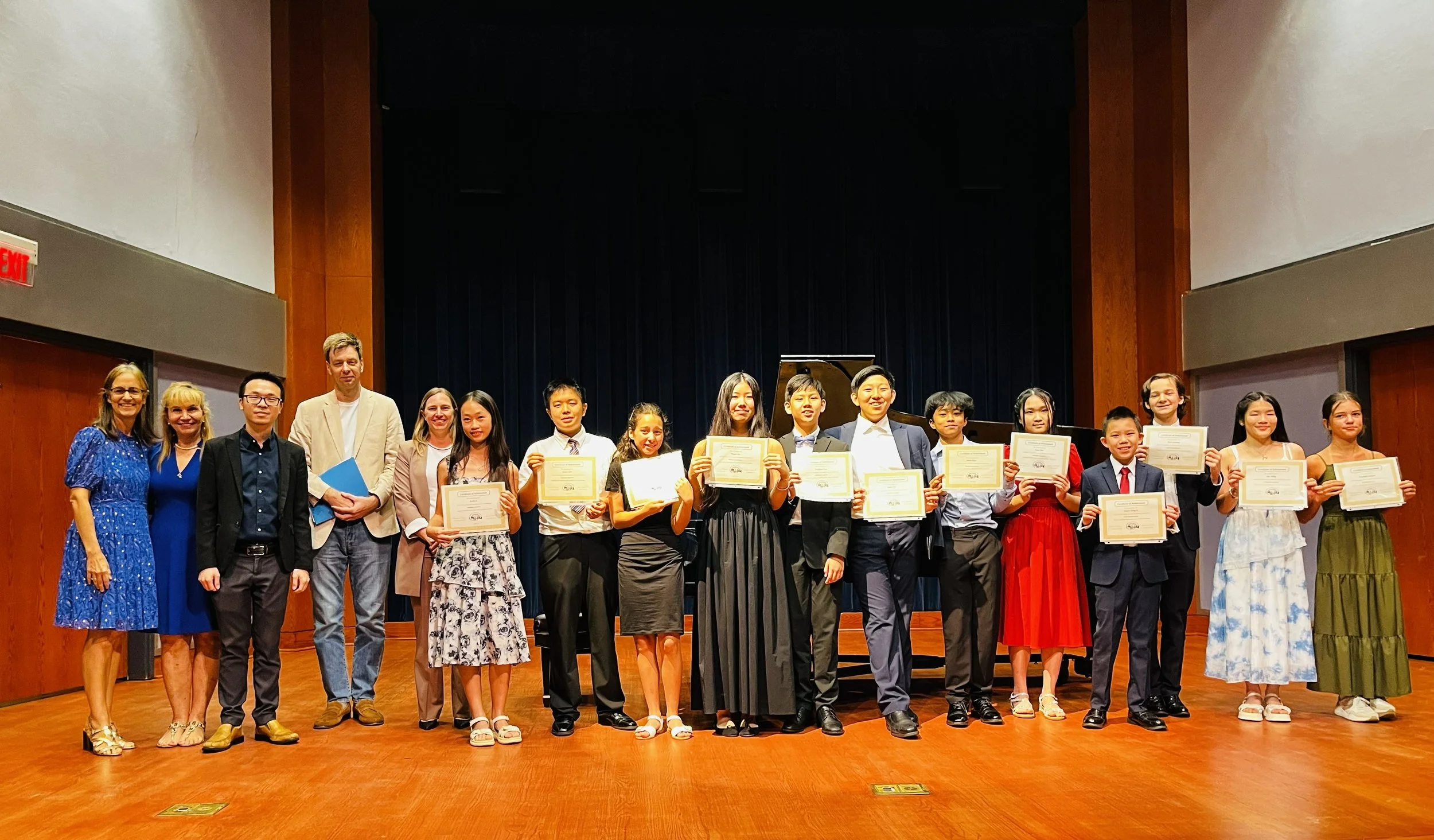 Group of children and adults on stage, holding certificates, in a wood-paneled auditorium, celebrating an achievement or graduation.