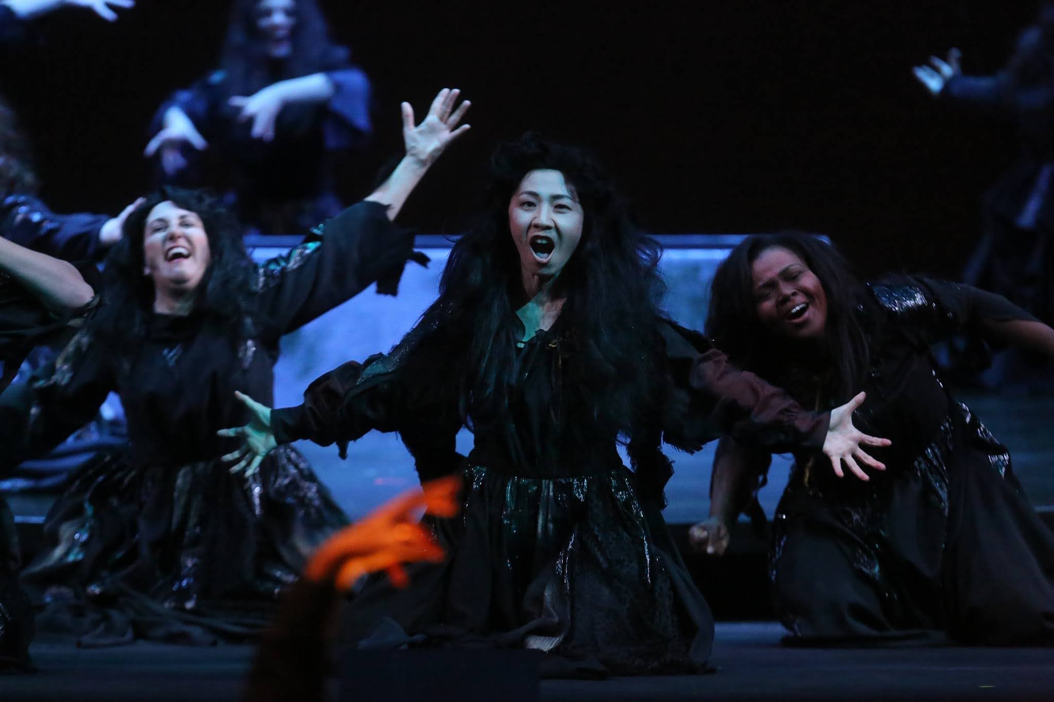 Nana Morimoto performing in an intense opera scene, dressed in black with long black hair, center stage, extending her arms in a dramatic moment during a theatrical production.