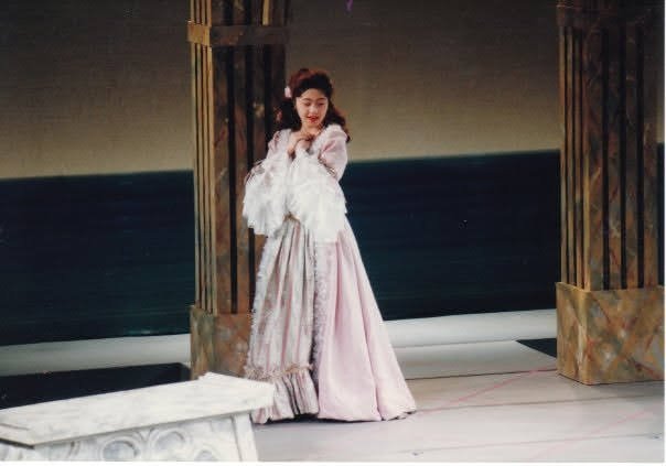 Nana Morimoto performing in an opera production, dressed in a historical costume on stage with wooden columns and a scenic backdrop of the sea and sky.