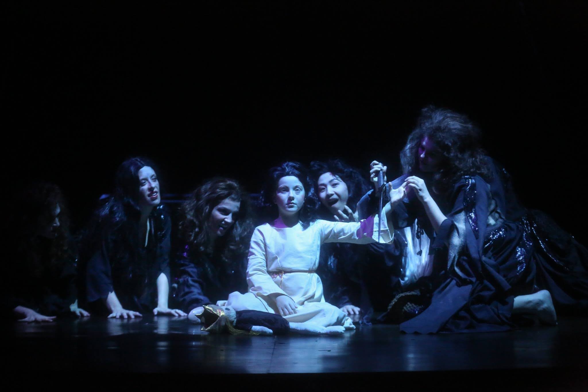 Theater performance with five women sitting on stage in dark costumes, appearing to act out a scene with the central woman in a white outfit holding an object, illuminated against a black background.