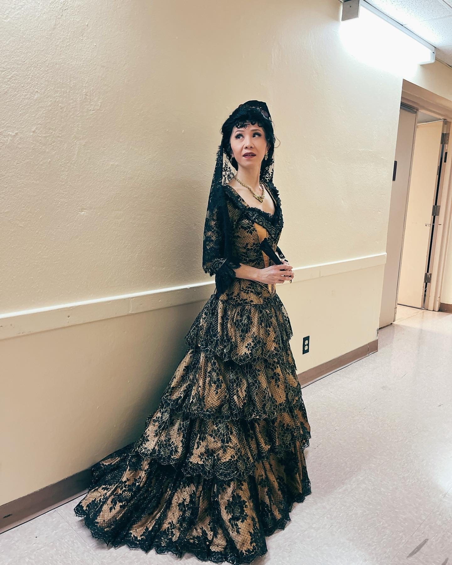 Nana Morimoto in a black and gold opera gown and headscarf, preparing for a classical vocal performance, photographed in the backstage hallway before an opera production.