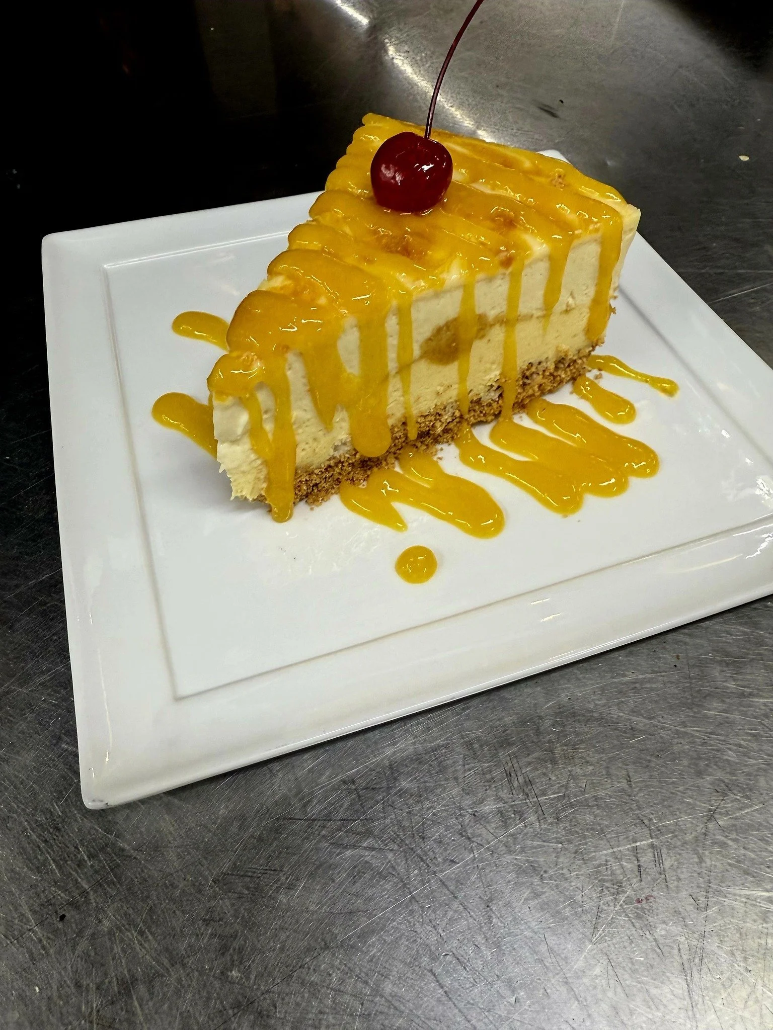 Slice of cheesecake with yellow caramel sauce and a cherry on top, served on a white square plate.