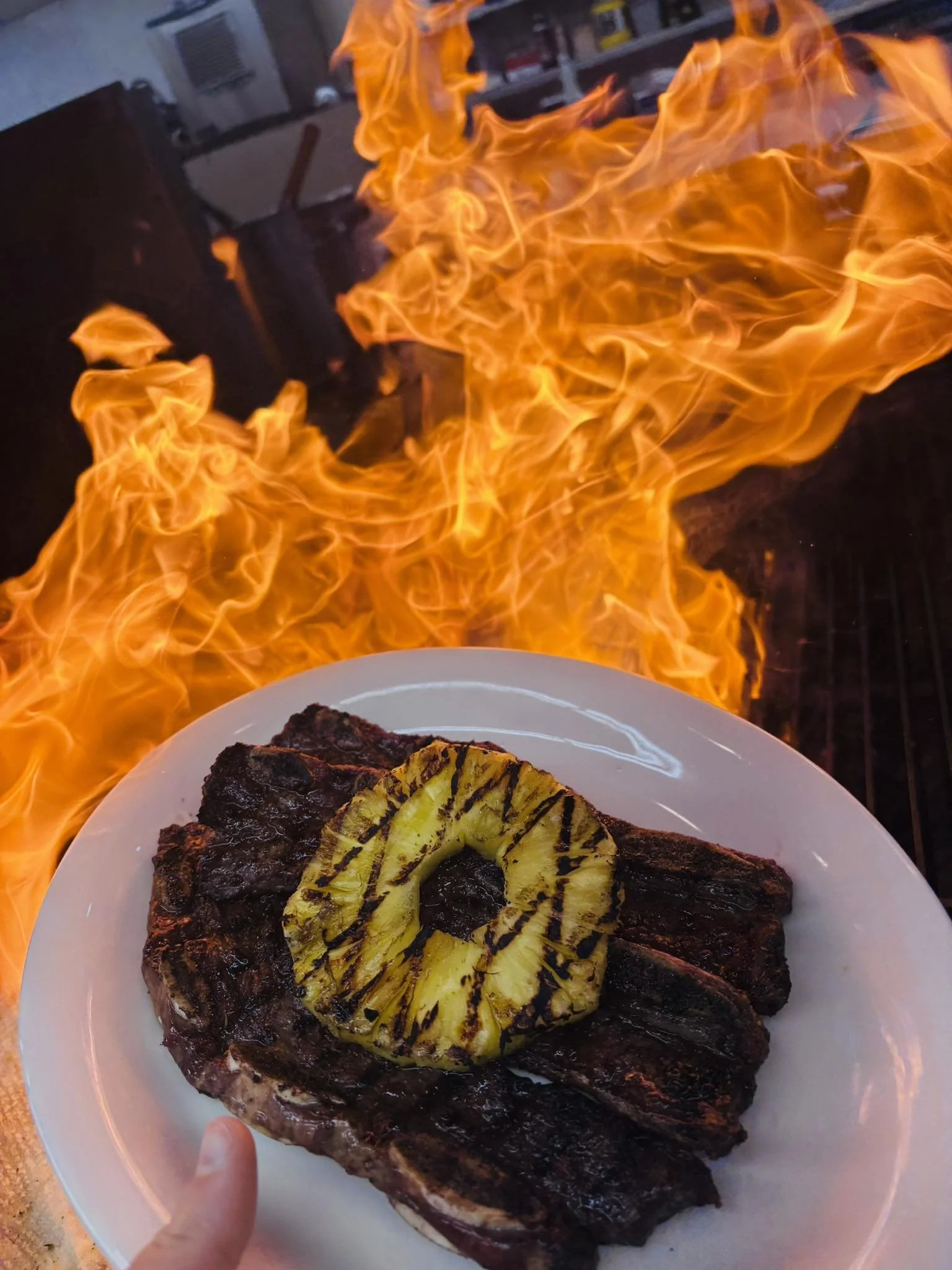 A plate of grilled steak with a grilled pineapple ring on top, held in front of a large open flame or grill fire.