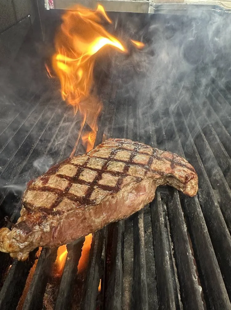 A thick cut of steak with grill marks is cooking on a grill over an open flame.