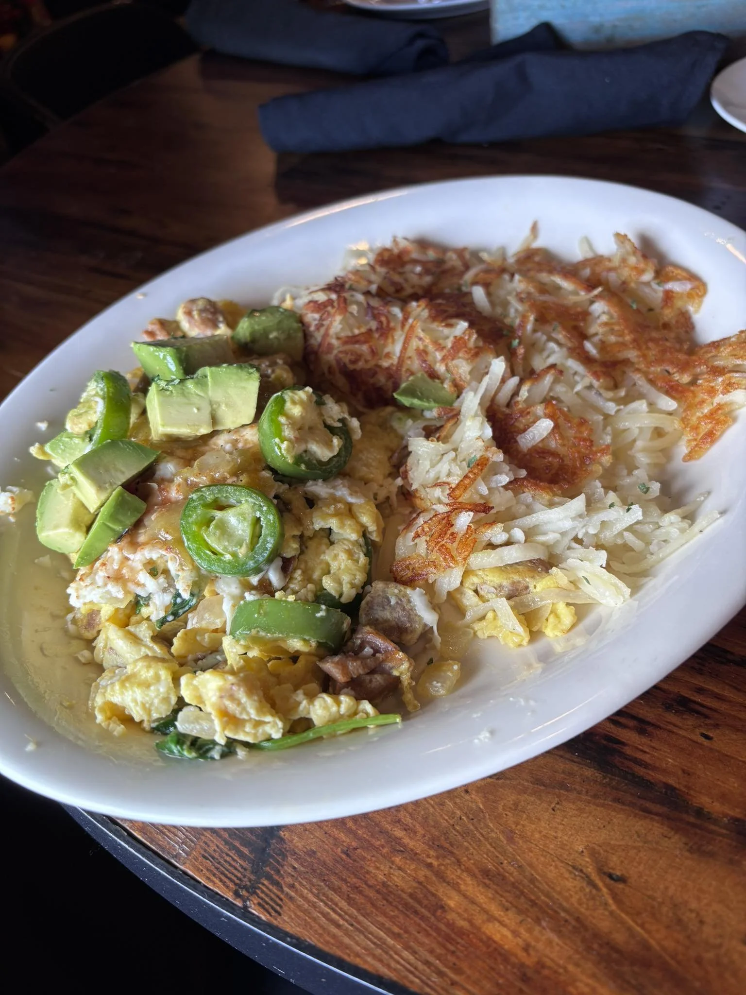 Breakfast plate with scrambled eggs, sliced avocado, jalapeño peppers, shredded cheese, and hash browns.