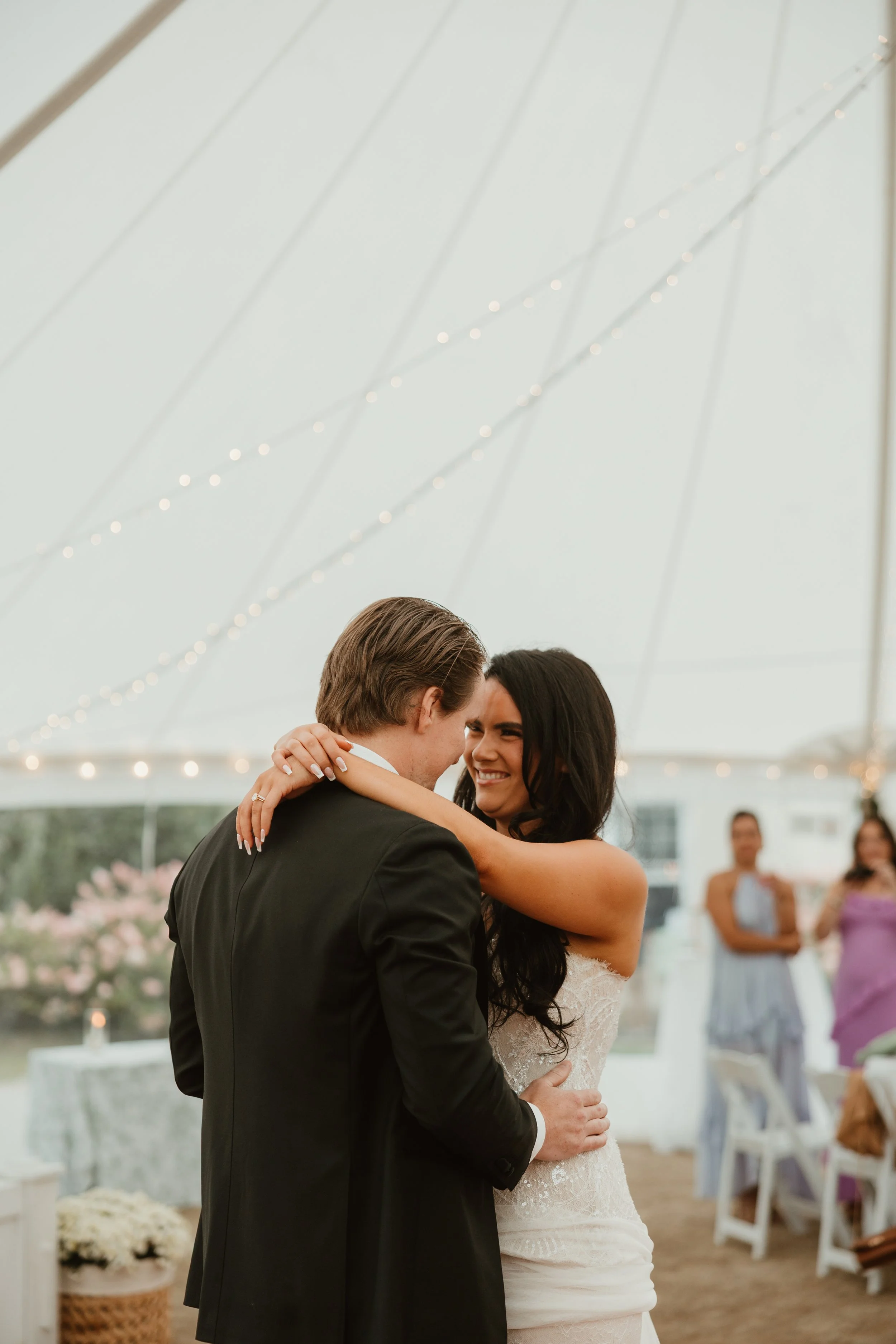 A couple sharing a dance at their wedding reception, under a white tent decorated with string lights, with guests watching in the background. This wedding was filmed by Shiang Studios.