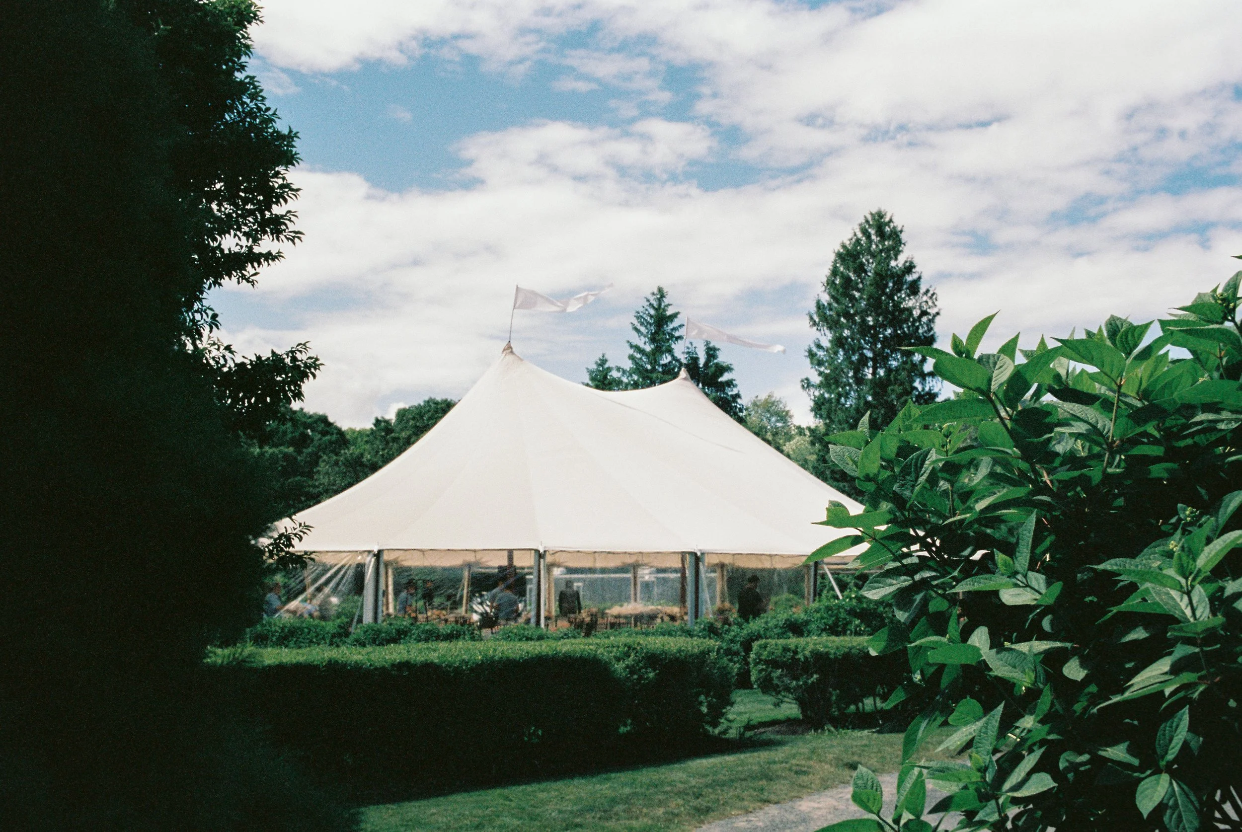 A large white event tent set up outdoors surrounded by lush green bushes and trees on a partly cloudy day. This wedding was filmed by Shiang Studios. This tent is located at the Connemara House Farm in Topsfield MA.