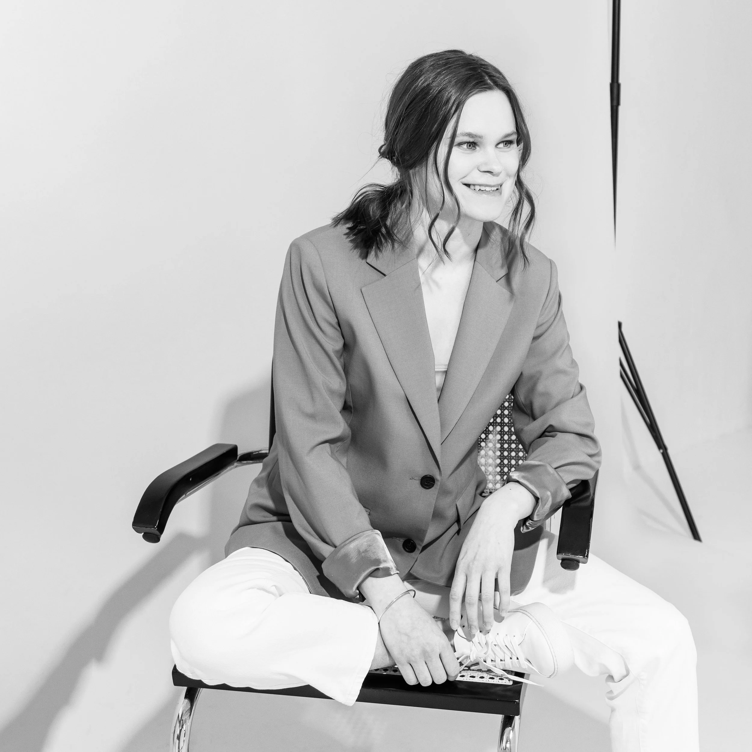 Black and white photo of a woman sitting on a chair, smiling, with her right leg crossed over her left, and tying her shoelace. She has shoulder-length wavy hair and is wearing a blazer and white pants.