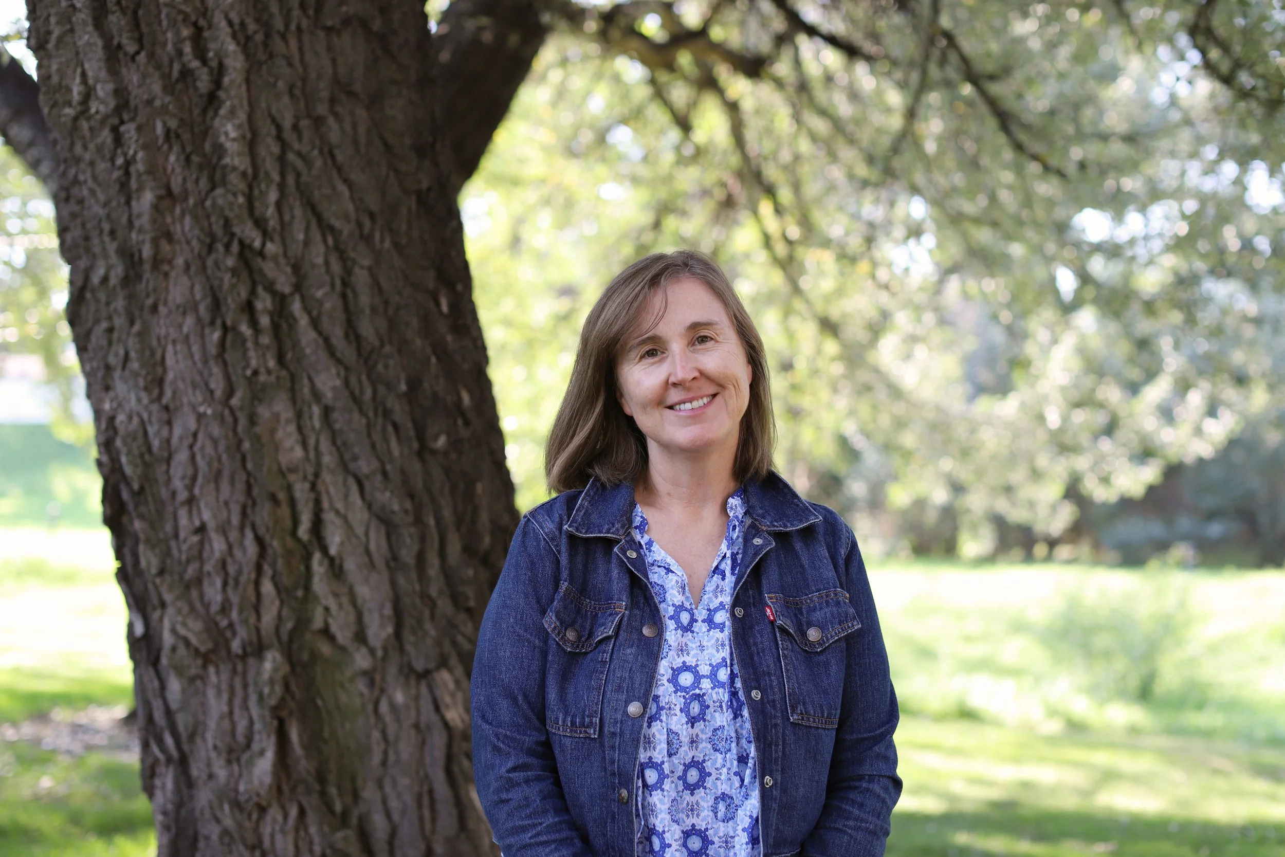 A woman with brown hair smiling outdoors next to a big tree in a park with green grass and trees in the background.