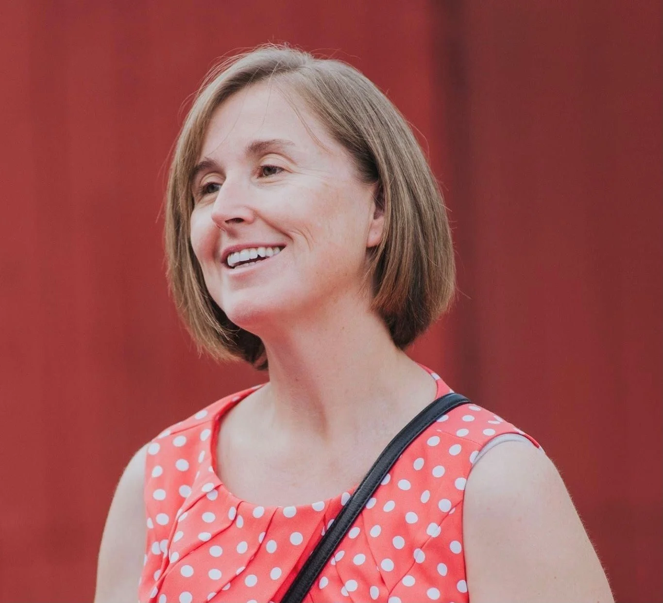A woman with short brown hair smiling, wearing a sleeveless coral dress with white polka dots, and carrying a black strap purse over her shoulder, standing against a red background.