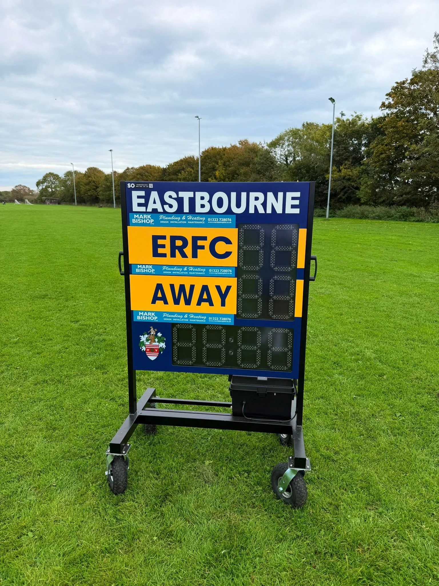 A scoreboard on a grassy sports field showing the team Eastbourne, sponsorship ads, and empty digital score displays.