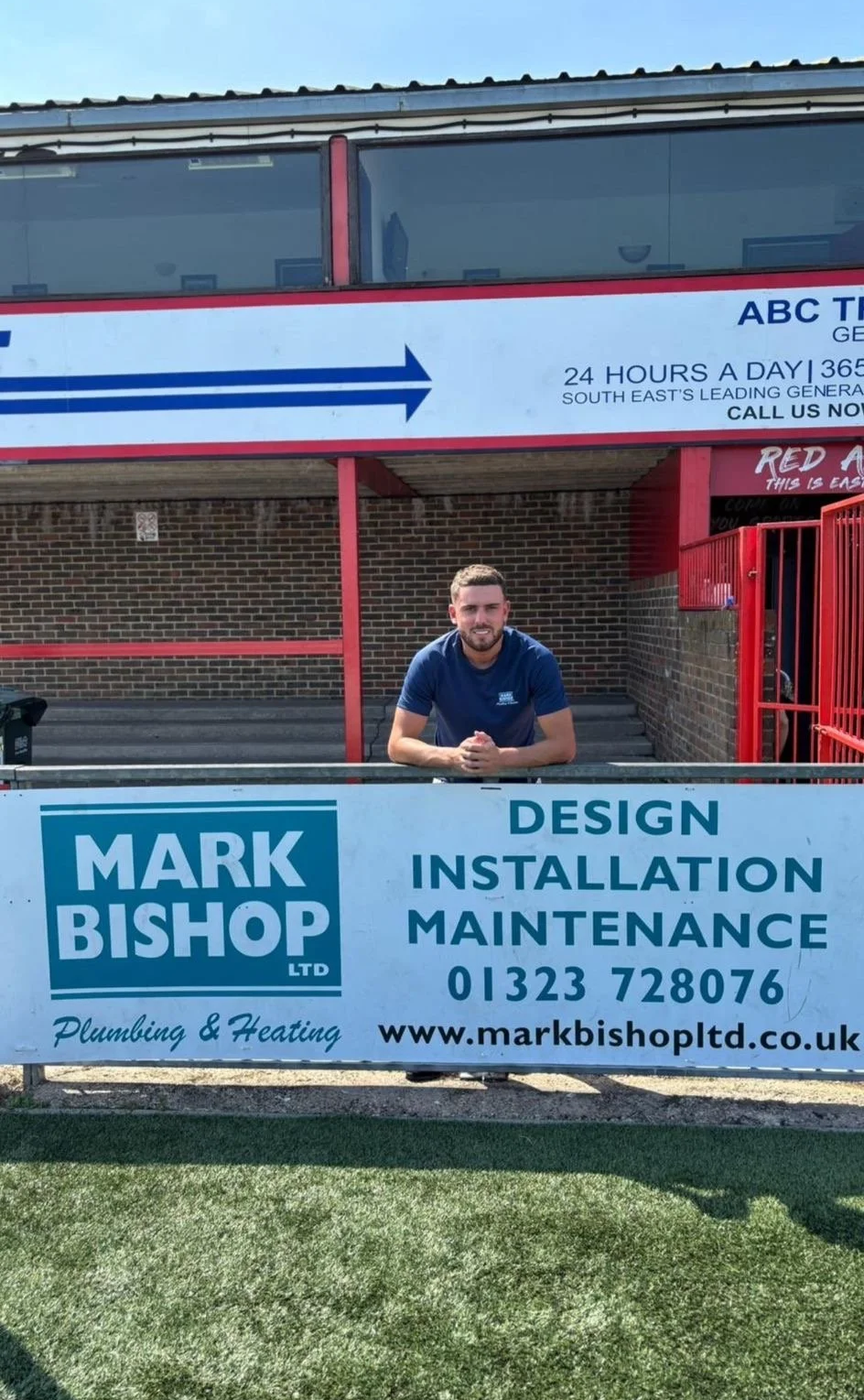 A man sitting behind a sign for Mark Bishop Ltd, a plumbing and heating company, with information about their services and contact details, outside a building with a brick wall and a staircase. Sponsorship for eastbourne borough football club