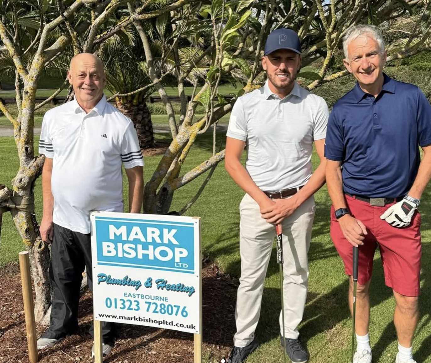Three men standing on a golf course, with a sign for Mark Bishop Ltd Plumbing & Heating in Eastbourne, and trees in the background. Willingdon Golf Course, eastbourne, east sussex sponsorship