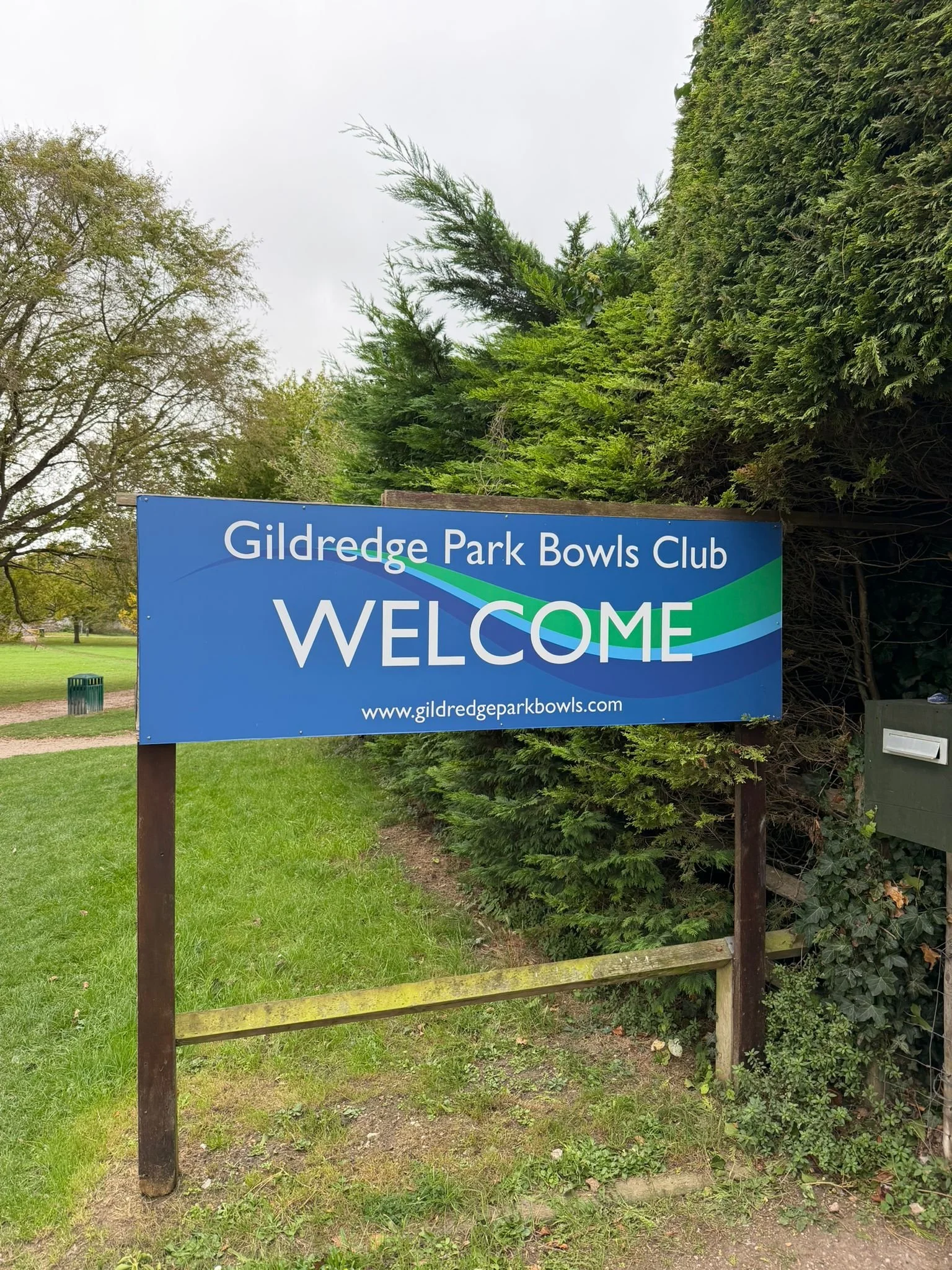 A bocce ball, a white ball, and a metal bocce ball on grass at a bocce court, with the logo of Gildredge Park Bowls Club overlaid.
