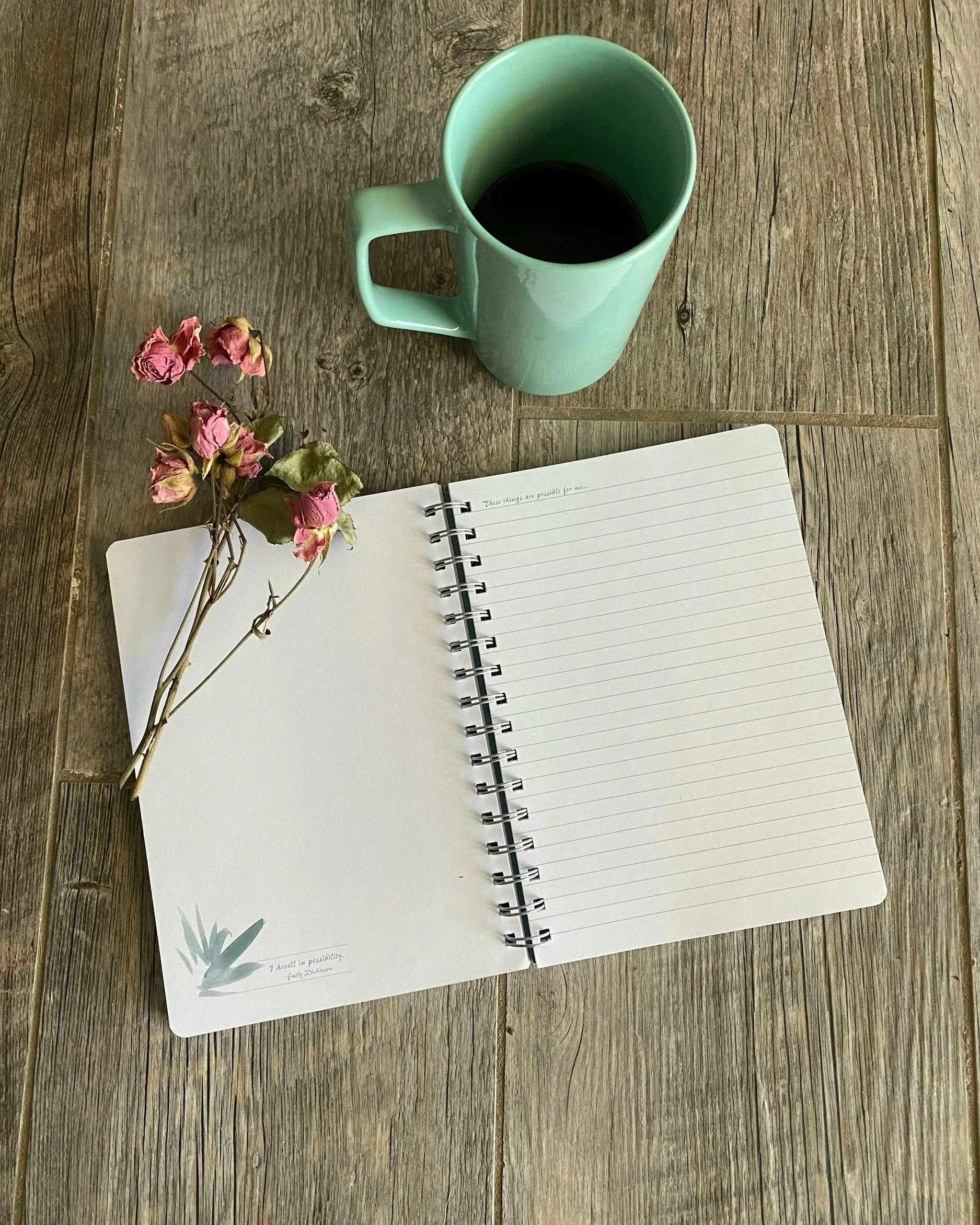 Open lined journal on wooden surface with pink dried flowers, a teal coffee mug filled with dark coffee.