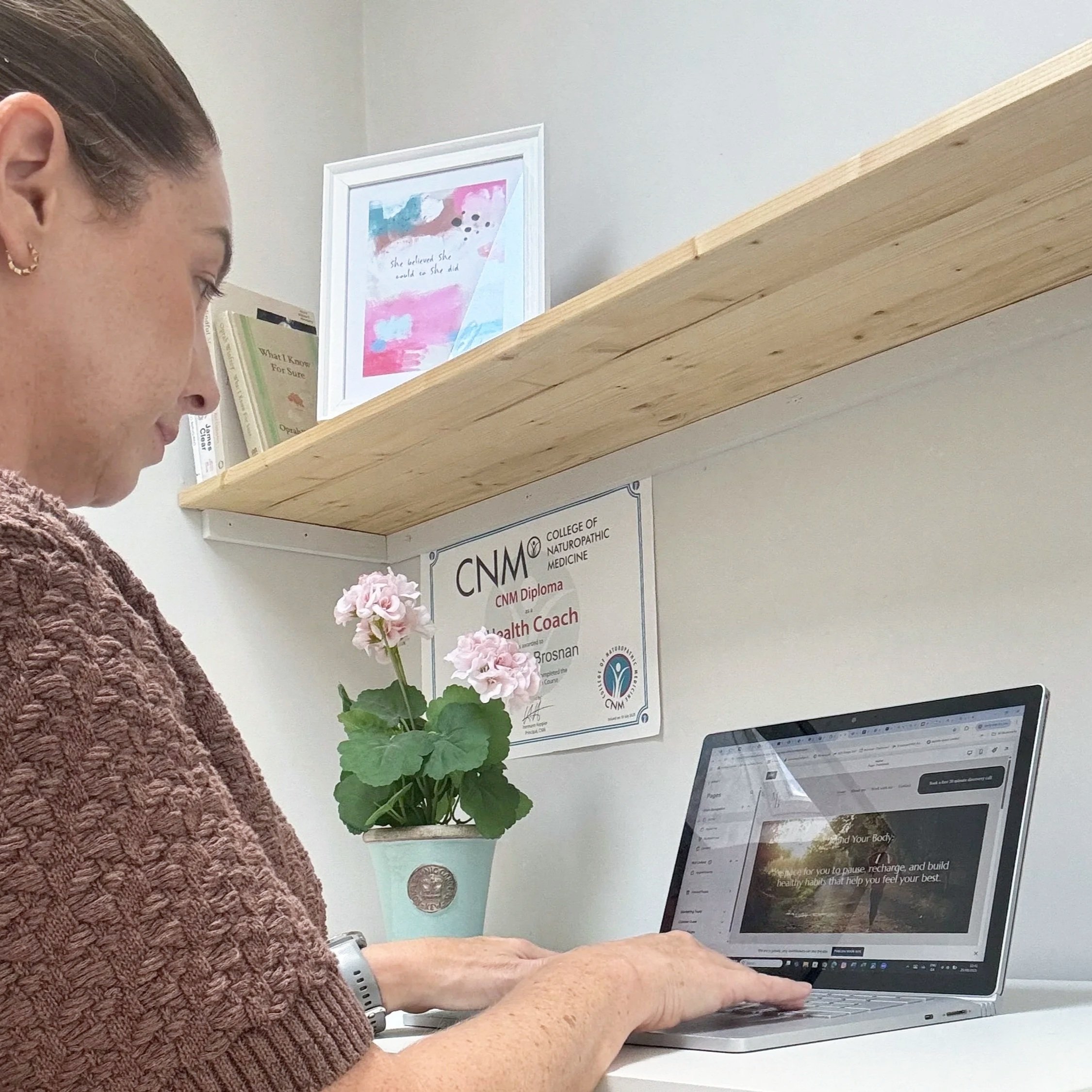 A person with short hair wearing a brown knit sweater and a white wristwatch working on a silver laptop at a white desk. On the wall behind them, there are framed posters and books on a floating wooden shelf. The posters mention the college of neurosurgical and neurotrauma medicine, a diploma, and health coaching. The person's focus is on the laptop screen.