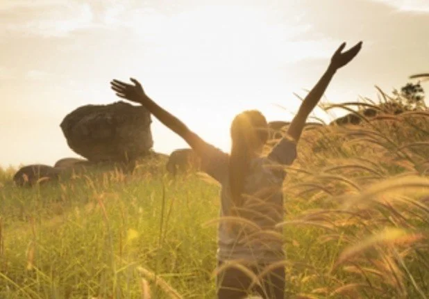 A person with long hair standing in a grassy field with arms raised, sunlight shining behind them, and large rocks in the background.