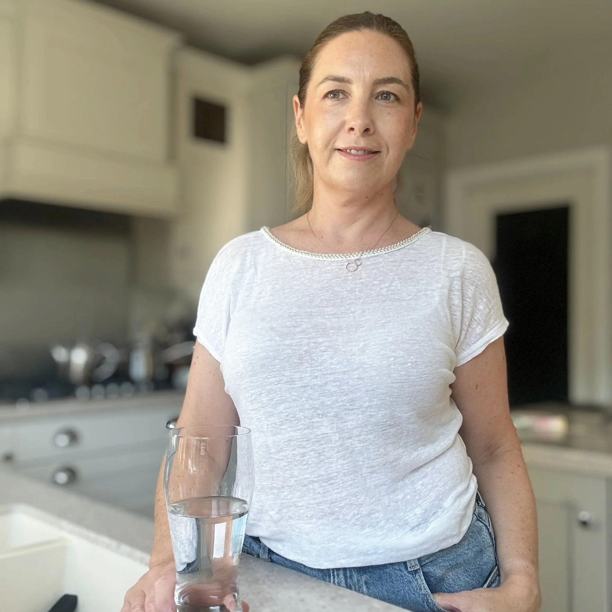 A woman in a white t-shirt holding a glass of water in a kitchen.