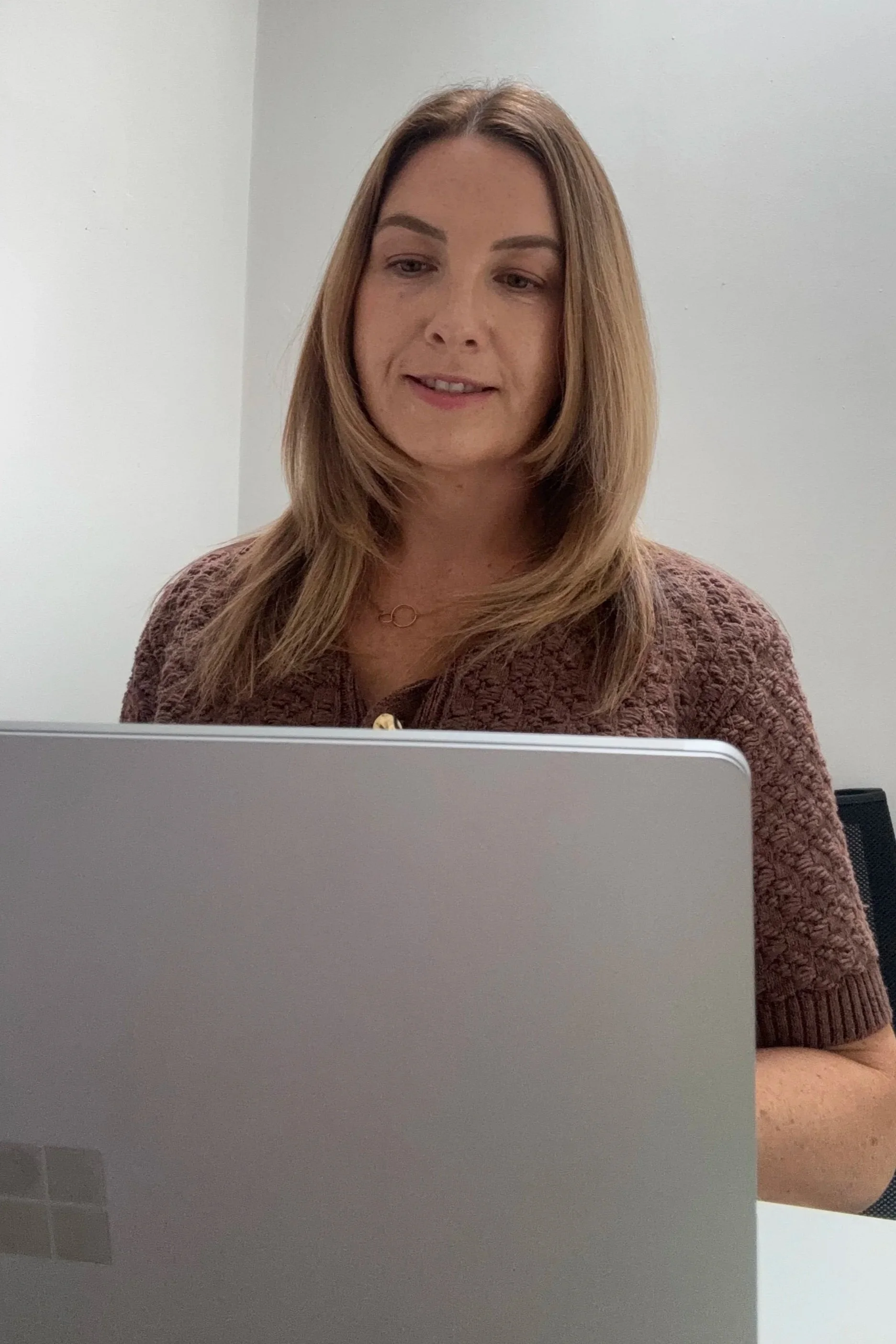 A woman with shoulder-length light brown hair, wearing a maroon knit sweater, looking at a silver laptop in a plain room.