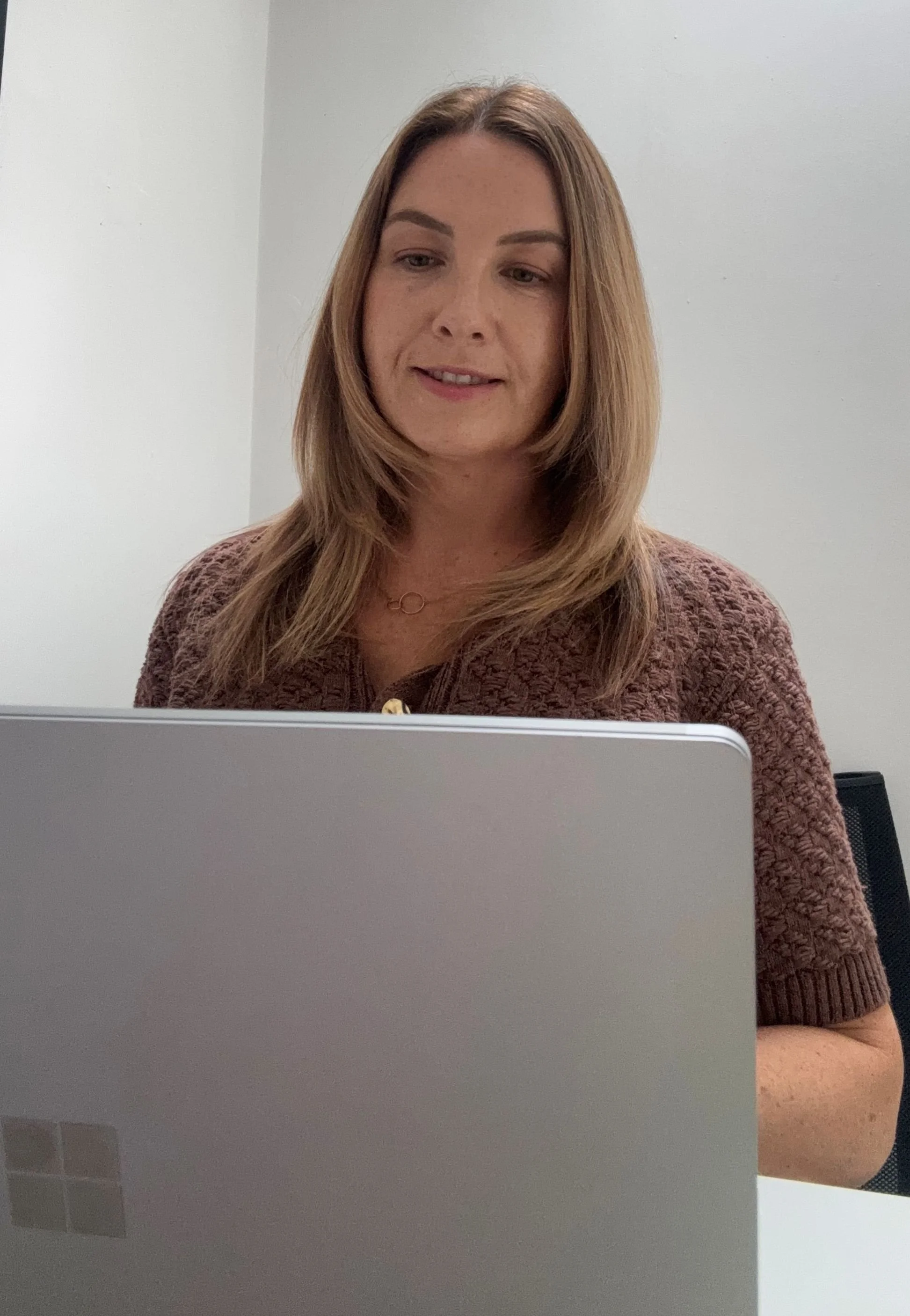 A woman with shoulder-length brown hair working on a silver laptop in a white-walled room.