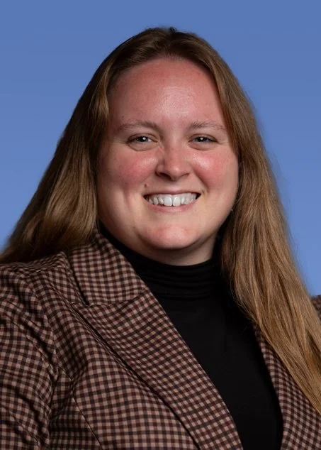 A young woman with long red hair, smiling, wearing a black top and a gray jacket, posed against a blue background.