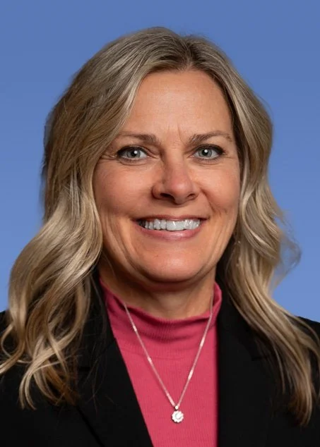A woman with shoulder-length blonde hair smiling, wearing a green blouse, silver necklace, and earrings, against a blue background.