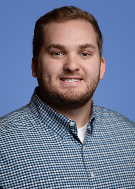 A young man with short blonde hair, a beard, wearing a navy blue polo shirt, sitting against a solid blue background.