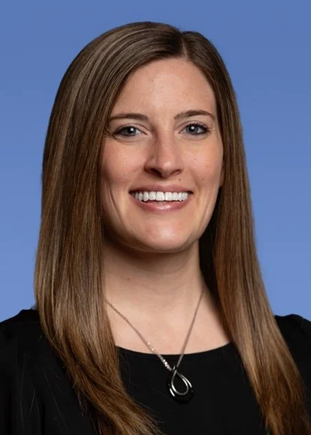 Professional portrait of a young woman with long, wavy blonde hair, wearing earrings and a black and white patterned blouse, smiling against a blue background.
