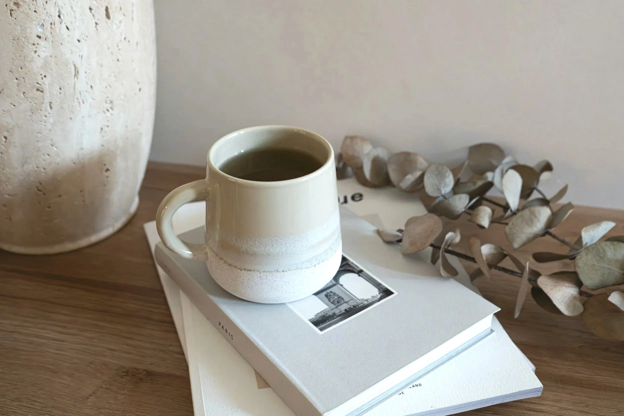 A white mug filled with dark coffee resting on a closed book, with a dried eucalyptus branch nearby on a wooden surface.