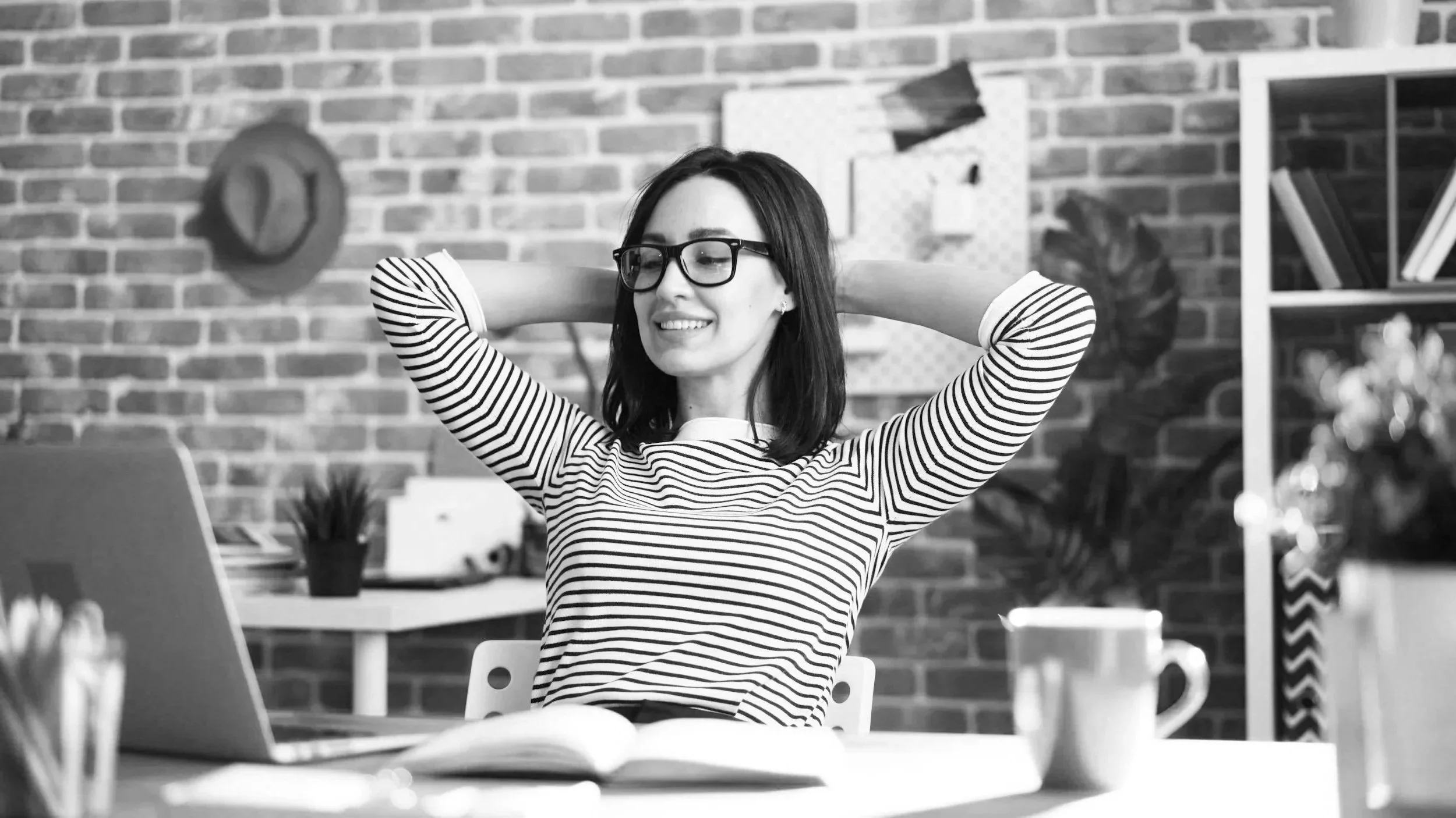 A woman with glasses and shoulder-length hair is sitting at a desk, smiling with her arms behind her head, in a room with a brick wall background.