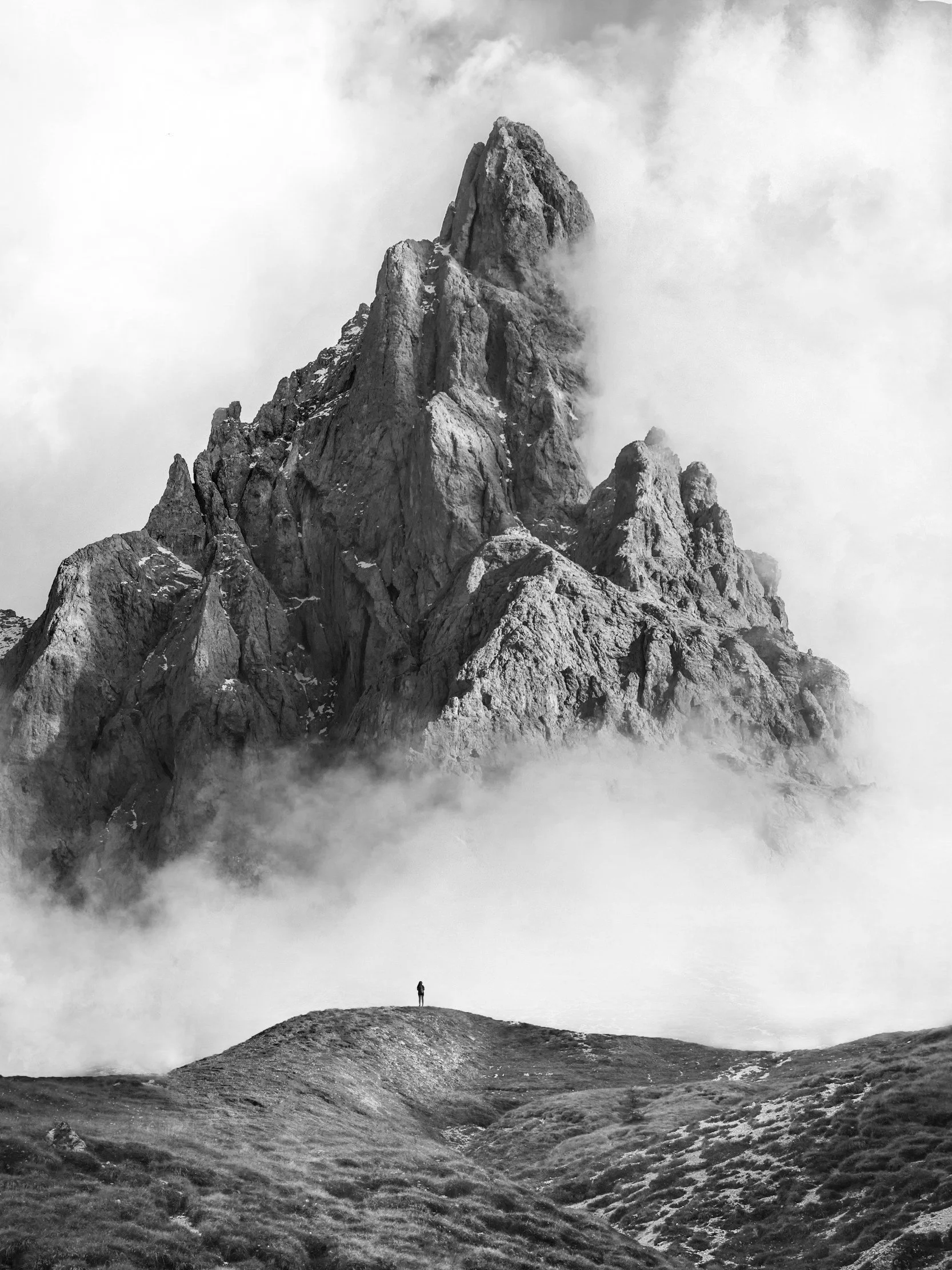 A person standing on a grassy hill beneath a tall, jagged mountain peak surrounded by clouds.