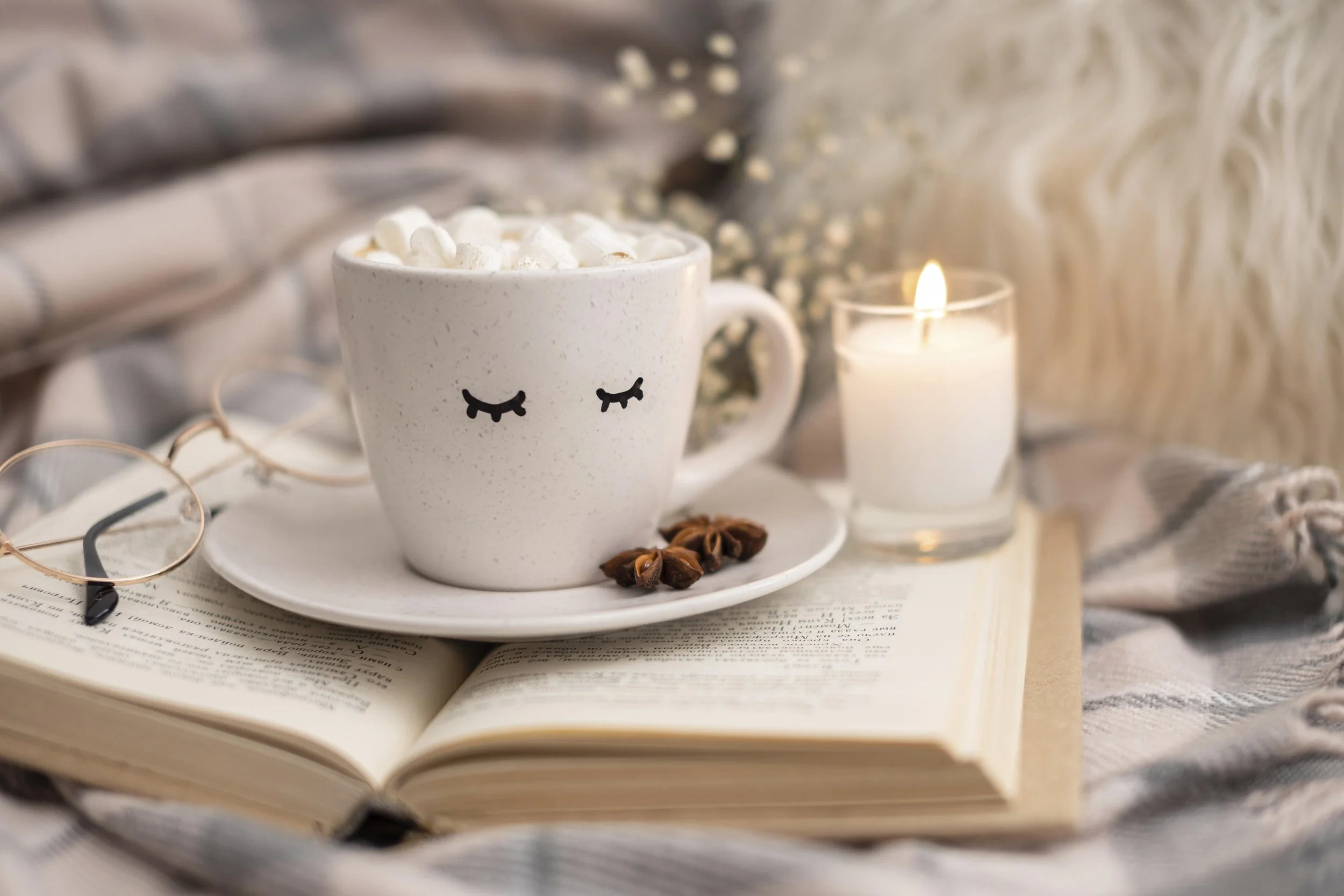 A cozy scene featuring an open book, a white mug with closed eye illustrations filled with marshmallows, a small plate with star anise, a lit candle in a glass holder, reading glasses, and a soft blanket or fabric in the background.