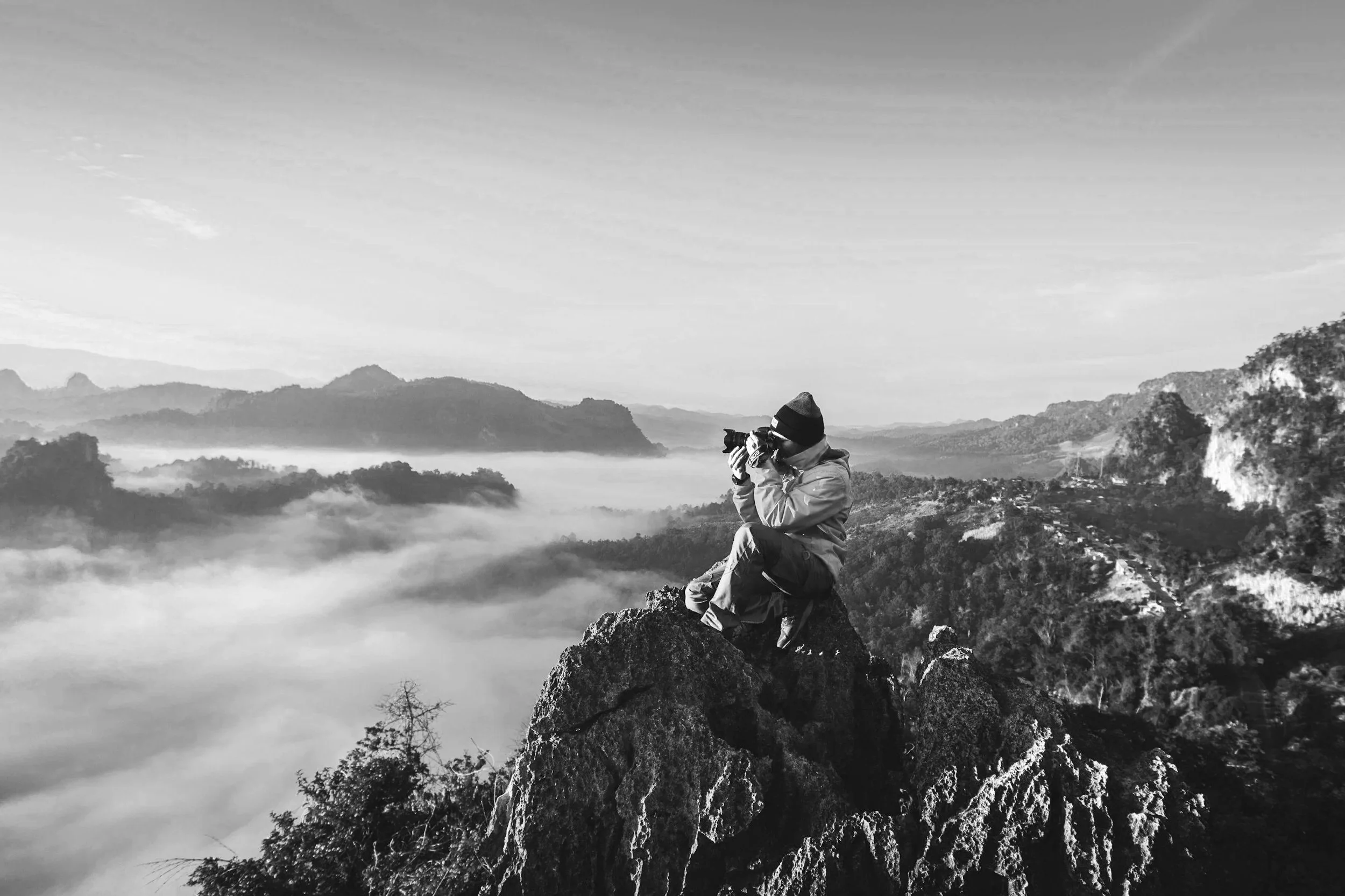 Person sitting on a rocky mountain peak, taking a photo with a camera, overlooking a foggy valley with mountains in the background.