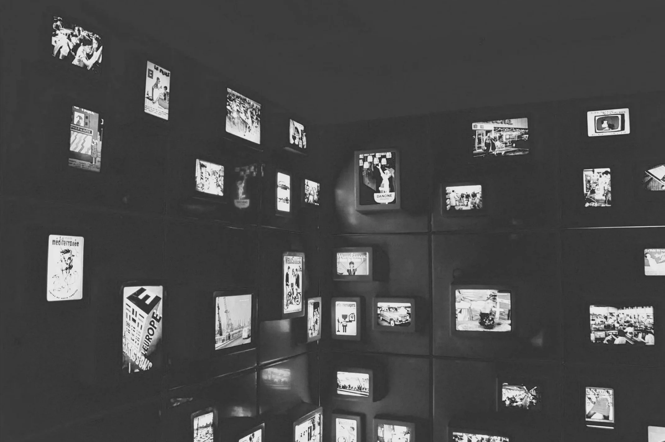 Wall with multiple small lit-up screens displaying various images and text in a dark room.