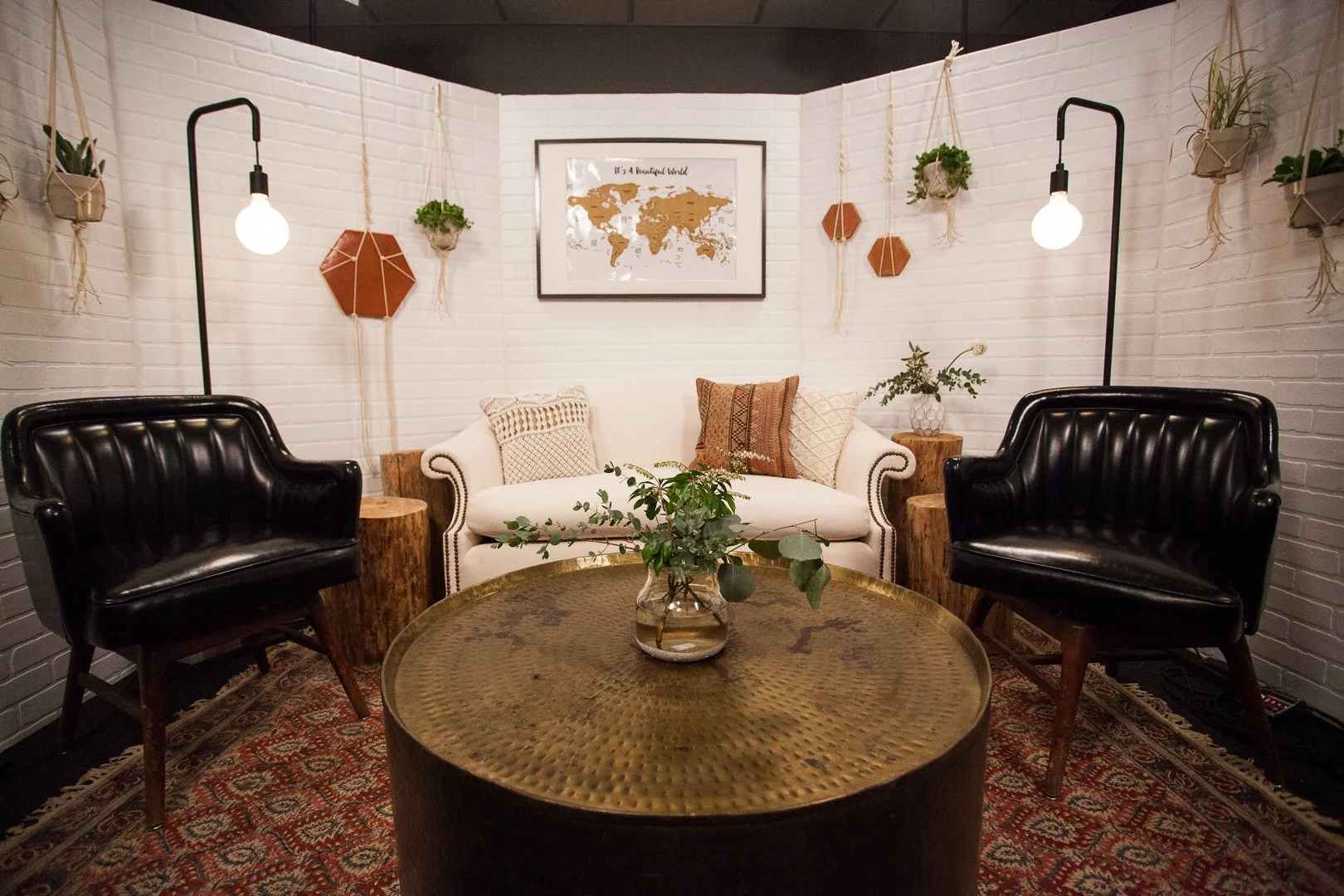 Cozy living room with white brick walls, two black leather armchairs, a white sofa with decorative pillows, a round wooden coffee table with a plant, and wall decorations including hanging plants, wooden wall art, and a world map art piece.