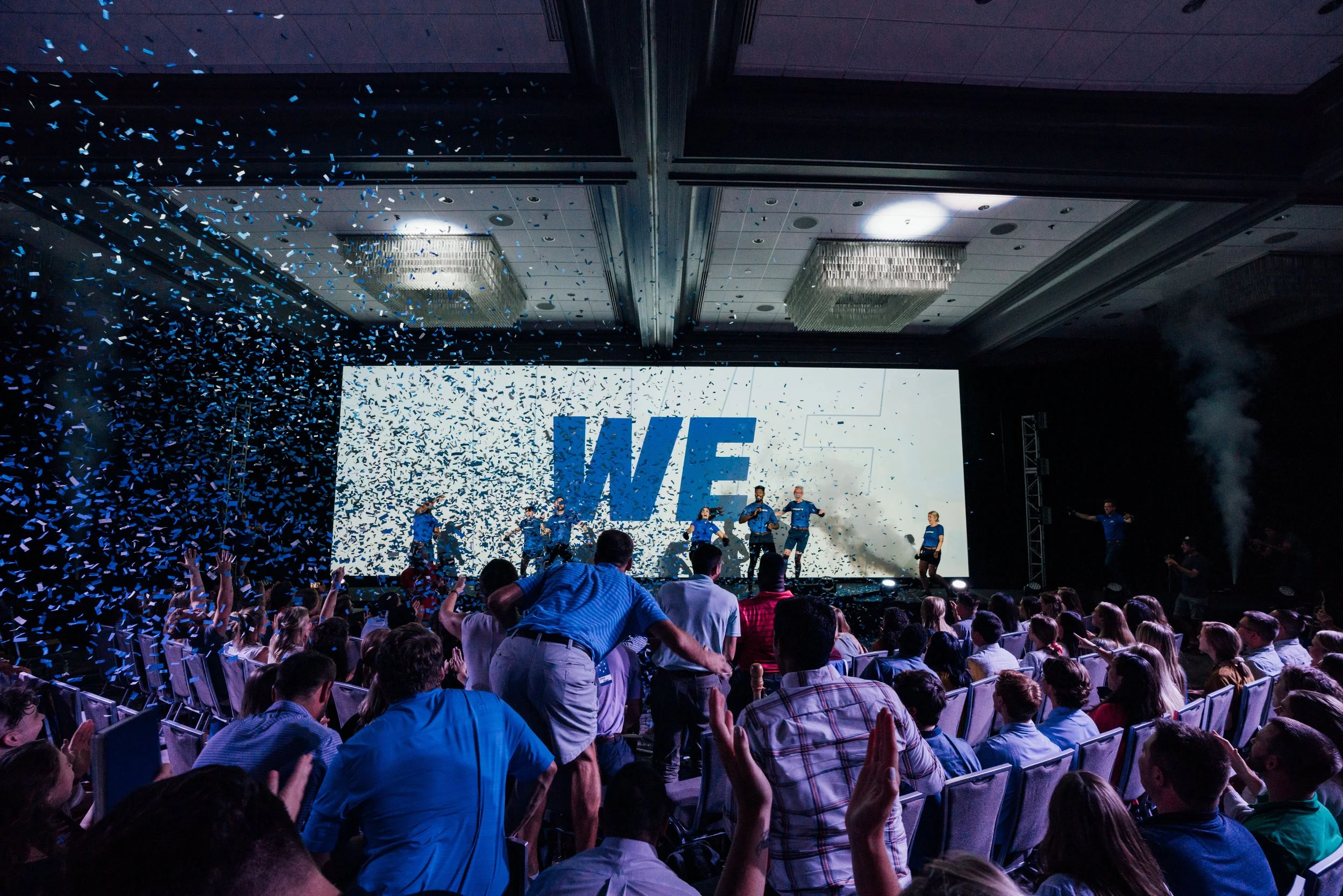 People attending a conference or event in a large hall with a stage, confetti falling, and a large screen displaying the words "WE" with performers on stage.