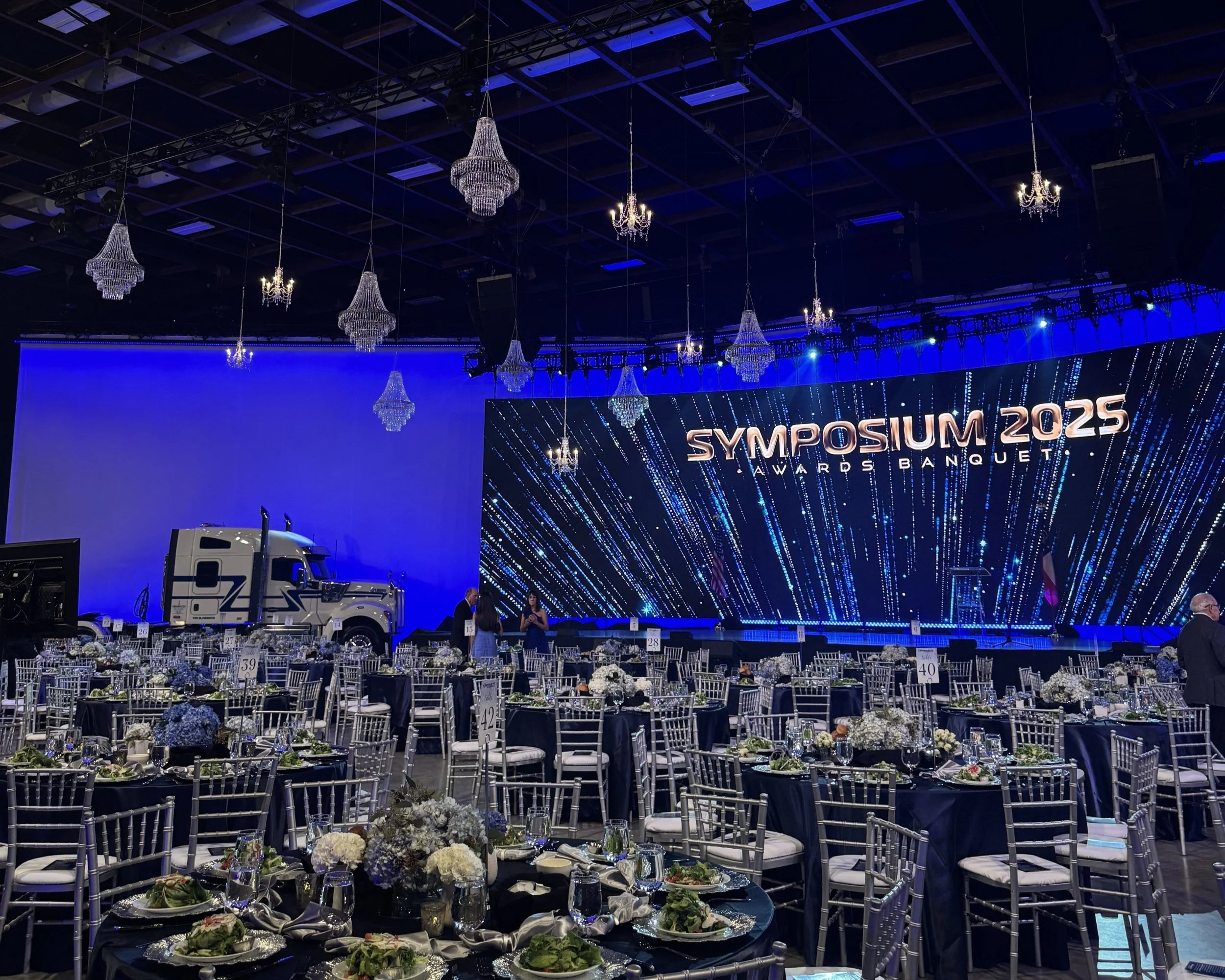 banquet hall set up with tables and chairs, decorated with floral centerpieces, prepared for a formal event. A large stage displays a sign reading 'Symposium 2025 Awards Banquet,' with chandeliers hanging from the ceiling and a truck positioned near