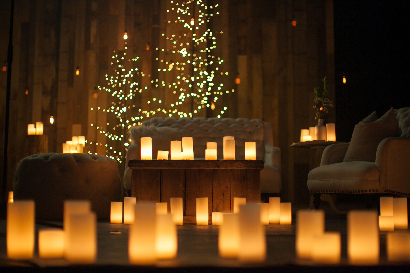 Cozy living room decorated with numerous flickering candles on tables and the floor, a tufted beige sofa, upholstered armchairs, a wooden coffee table, and a wall decorated with a string of small, warm white lights, creating a warm, ambient atmospher