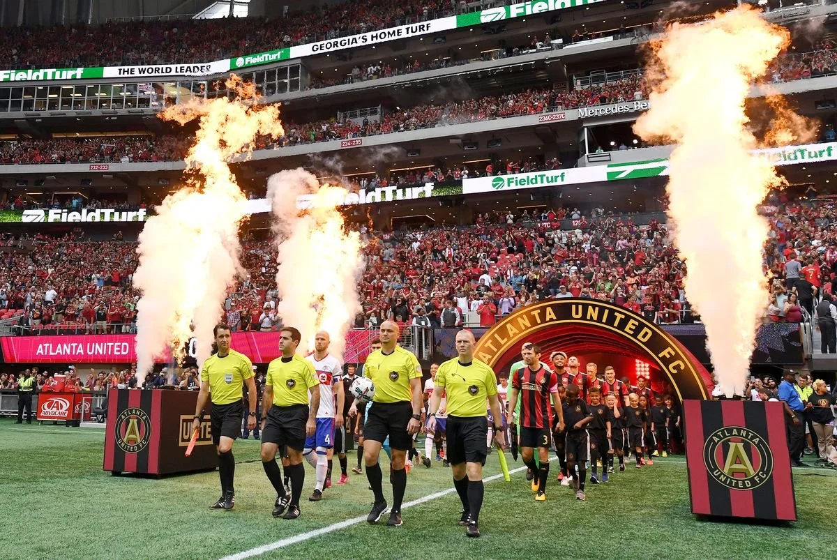 Soccer players and referees walking onto the field through an arch with flames and smoke, with a large crowd in a stadium in the background.