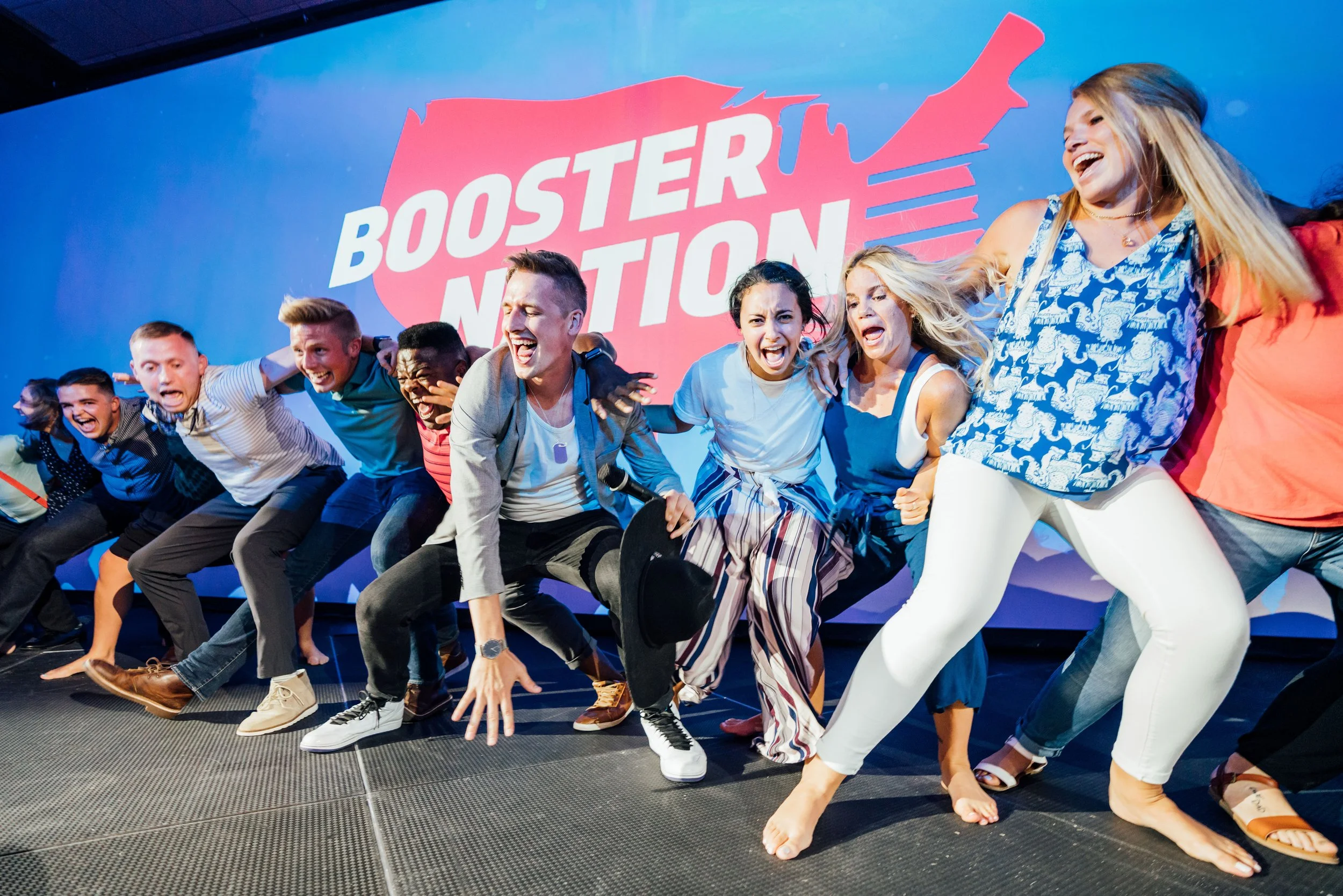 Group of people on stage participating in a team-building activity, laughing and leaning back in a tug-of-war style game, with a large sign in the background that reads 'Booster Nation'.