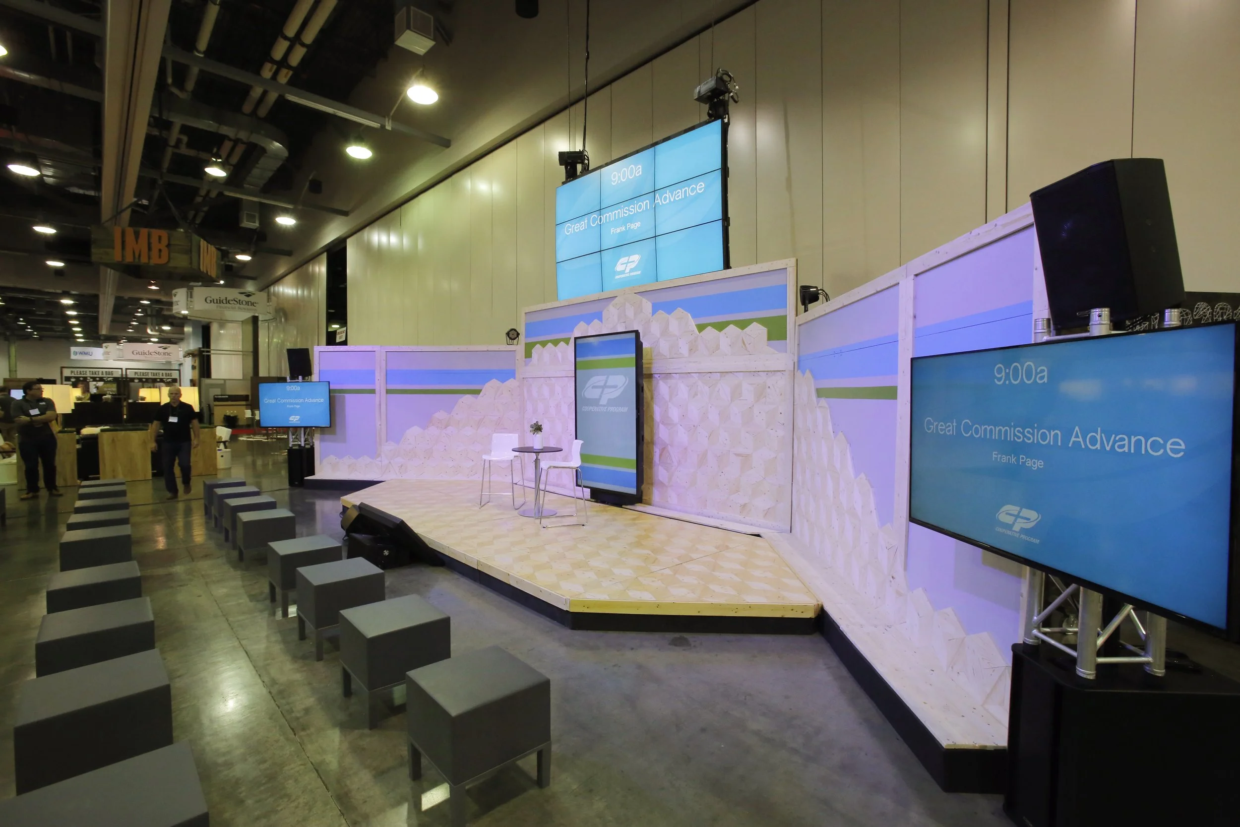 A stage setup at a conference with a large screen displaying 'Great Commission Advance' and the name Frank Page, with smaller screens and black chairs in front.